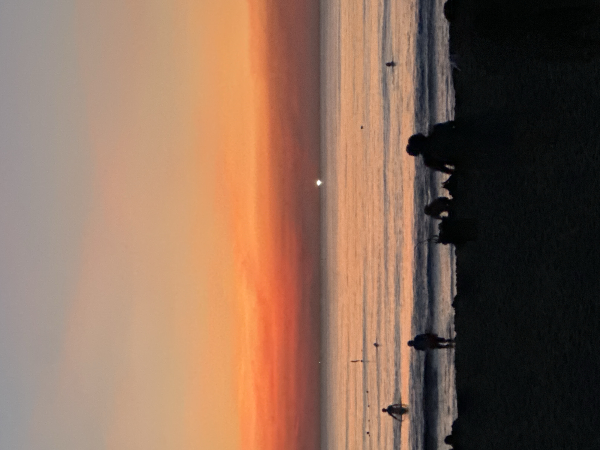 The image captures a serene beach scene at dusk in Da Nang, Vietnam. The sky is a gradient of soft grays and warm oranges, suggesting the sun has recently set or is about to rise, casting a gentle glow over the water. The ocean, a deep blue with streaks of orange reflection, is calm, with small waves lapping at the shore.
Silhouettes of people are visible along the shoreline and in the shallow water. One figure stands near the water's edge, facing away from the camera, while others are scattered further out or near the dark, silhouetted beach. It appears to be a leisurely evening, with individuals enjoying the tranquil atmosphere. The overall mood is peaceful and reflective. There is no visible text in the image.