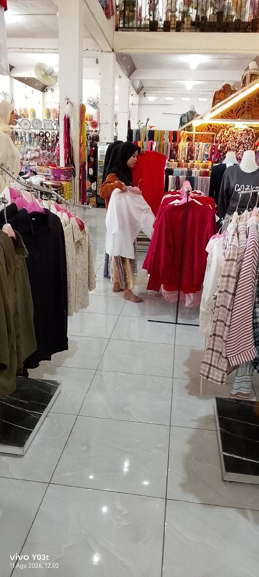 The image depicts an indoor retail environment, likely a clothing store, with tiled flooring. In the foreground, clothing racks display various garments, including shirts and patterned fabrics. The central focus is a woman wearing a hijab and a long-sleeved top, holding a white garment. She is standing barefoot on the floor, seemingly examining the clothing. Behind her, more clothing is visible on racks and mannequins, with a red garment prominently displayed on a stand. The background shows more shelving and displays of merchandise, suggesting a busy marketplace or shop. The timestamp in the lower-left corner indicates "vivo Y03t 11 Agu 2026, 12.02", but this is likely metadata from the device and not directly relevant to the scene itself, though it suggests the photo was taken in Atula, Indonesia.