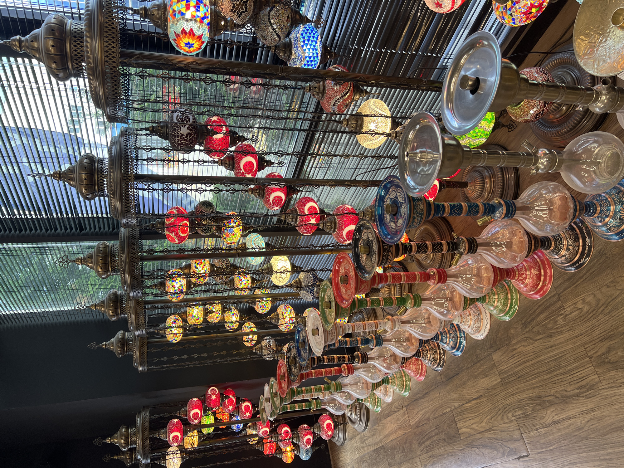 The image captures an interior scene, likely a shop, market stall, or a themed lounge in Krasnodar, Russia, featuring a vibrant and extensive display of ornate decorative items. The primary subjects are numerous colorful lamps and a collection of shisha pipes.
On the left and extending upwards, multiple rows of traditional mosaic lamps are suspended. These spherical and teardrop-shaped lamps are crafted from small, intricately arranged pieces of colored glass, creating dazzling patterns in hues of red, blue, green, yellow, and orange. Many lamps feature a crescent moon and star motif, often associated with Turkish culture. They are housed within decorative metal cages or frames, hanging from chains attached to a larger metal structure or shelving system.
To the right, a parallel display showcases a variety of shisha pipes or hookahs. These feature clear and colored glass bases, along with intricately decorated metal stems and trays. The bases exhibit diverse patterns and colors, including reds, greens, blues, and gold, with some featuring painted designs. These items are also arranged in orderly rows, suggesting either retail stock or decorative elements.
The background reveals a ceiling composed of horizontal slatted blinds, through which bright daylight filters, illuminating some of the objects and suggesting it is daytime. Outside the blinds, hints of foliage and buildings are visible. The foreground and bottom right of the image show a warm-toned wooden floor.
No people or discernible text are visible. The overall scene evokes a rich, exotic, and artisan atmosphere, highlighting the beauty and craftsmanship of the displayed items.