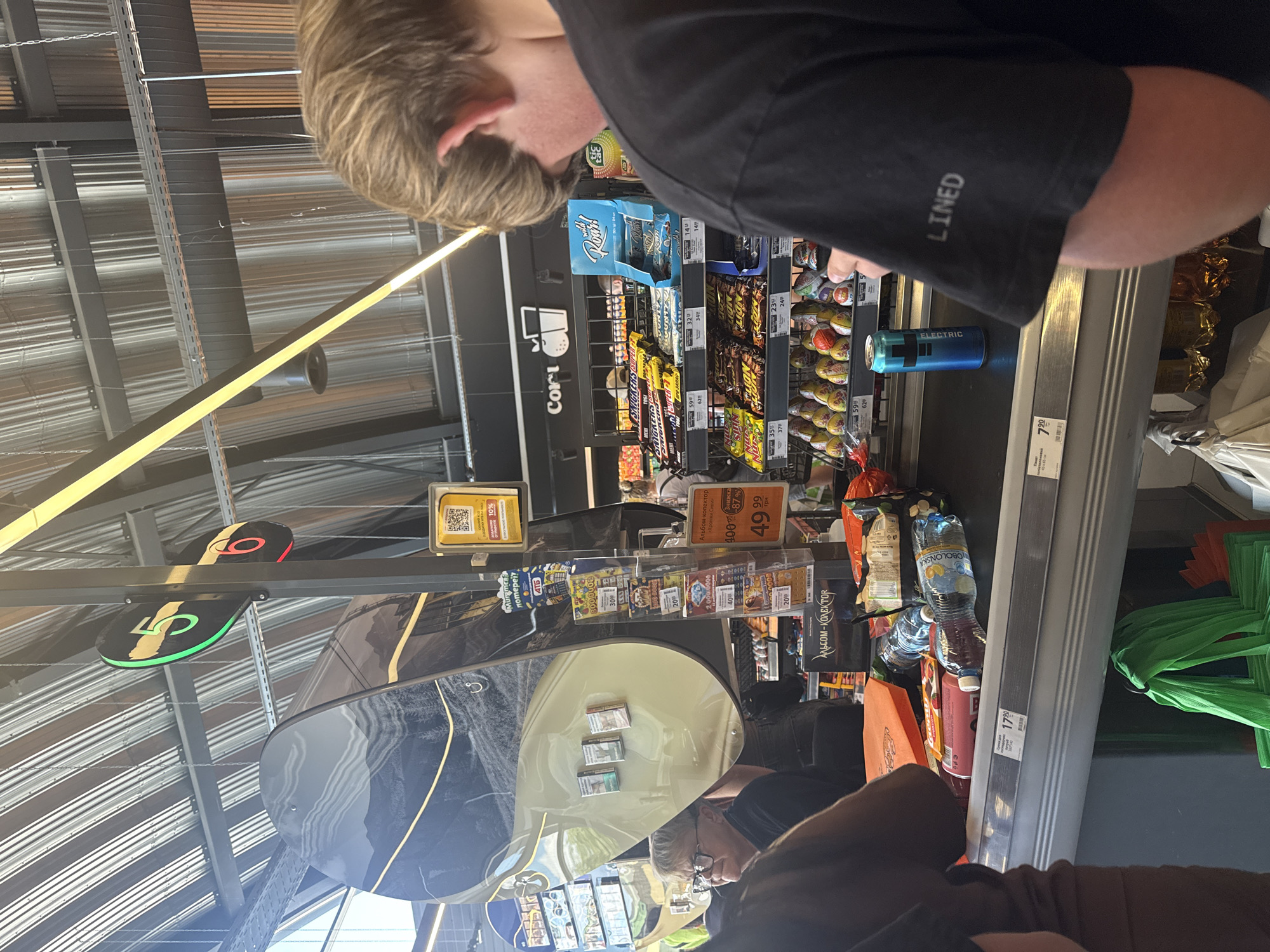 The image shows a checkout counter in what appears to be a supermarket in Dnipro, Ukraine, likely a Silpo or ATB based on signage. A young person with light brown hair and wearing a black t-shirt with the word "LINED" printed on the sleeve is standing at the counter, looking down at their hand and possibly the items being scanned. To the left, another person with glasses and dark hair is partially visible, looking towards the counter. The checkout belt displays several items, including water bottles, a pink tube-shaped item, and a blue can of "ELECTRIC" energy drink. Behind the counter, shelves are stocked with various snacks and confectionery, such as Snickers, Bounty, and Lion bars, along with what appear to be Easter eggs. A QR code is visible on a yellow sign, likely for promotions or payments. Overhead, the ceiling has a metallic, corrugated design, and signs indicating checkout lanes "5" and "6" are visible. Bags, including green and white reusable shopping bags, are hanging below the counter. The overall environment suggests a busy retail setting.