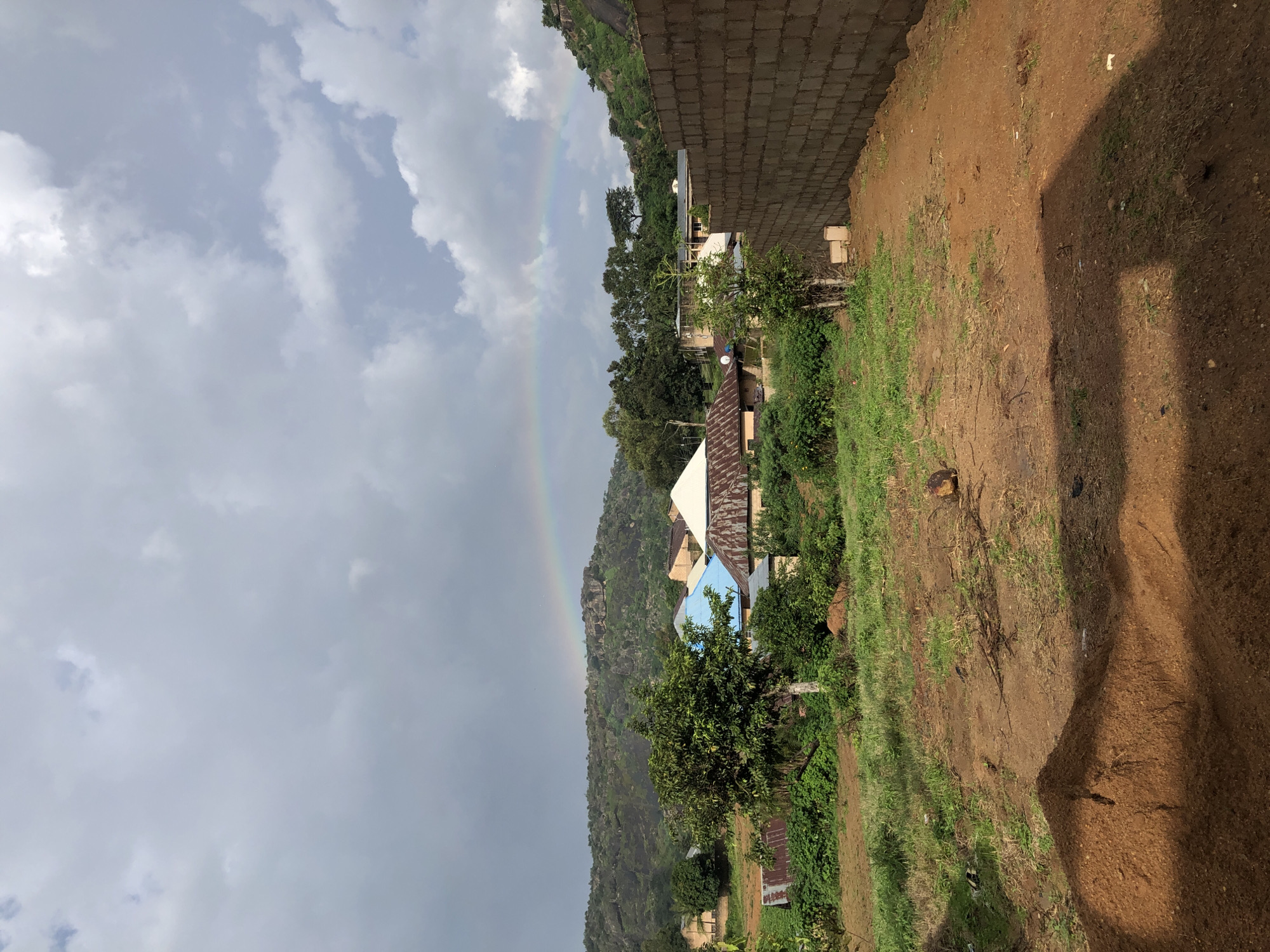 A pastoral landscape in Jos, Nigeria, is captured under a dramatic sky. A prominent rainbow arcs across the horizon, a vibrant contrast to the overcast clouds and the verdant hills in the background. The scene is dotted with a mix of traditional and more modern housing, characterized by corrugated metal and blue-tiled roofs. A partially constructed brick wall dominates the right foreground, suggesting ongoing development. The terrain is a mix of earth and sparse vegetation, with a pile of reddish-brown soil prominent in the lower left corner. The overall impression is one of rural life, with a touch of natural spectacle.