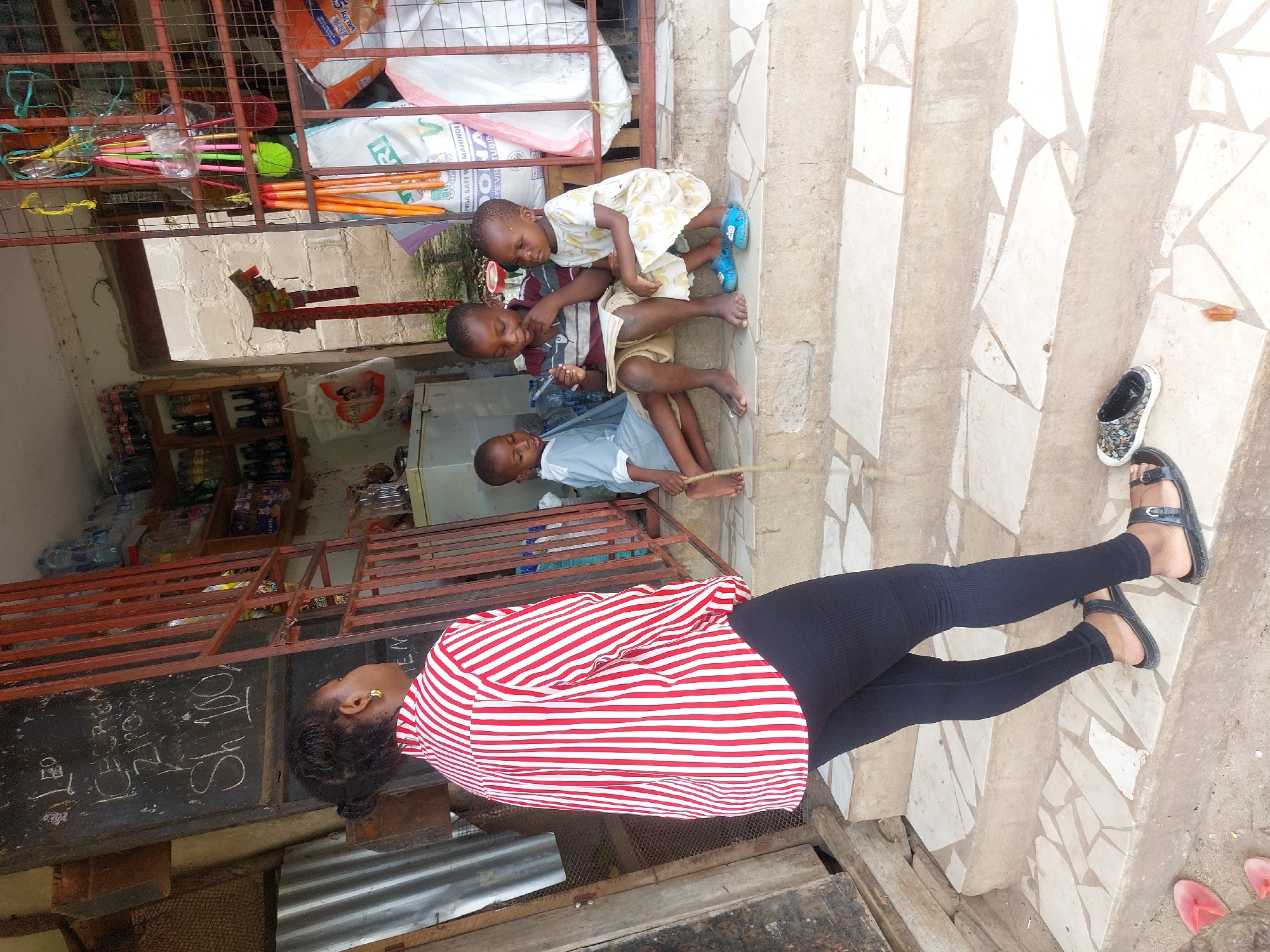 The image shows a scene in Dar es-Salaam, Tanzania. A woman wearing a red and white striped shirt and black leggings stands on a step next to what appears to be a small shop or stall. Her back is to the camera. In front of her on the steps sit three children, one in a light yellow dress, one in a maroon striped shirt and khaki shorts, and the third in a light blue shirt. The children are sitting close together and appear to be interacting. A pair of black sandals are next to her feet, and a pair of shoes are nearby. The shop has a metal rack with various items displayed, including bags, bottles, and other goods. There is a chalkboard with some writing on it near the woman. The steps and the surrounding ground are made of a mosaic tile pattern.