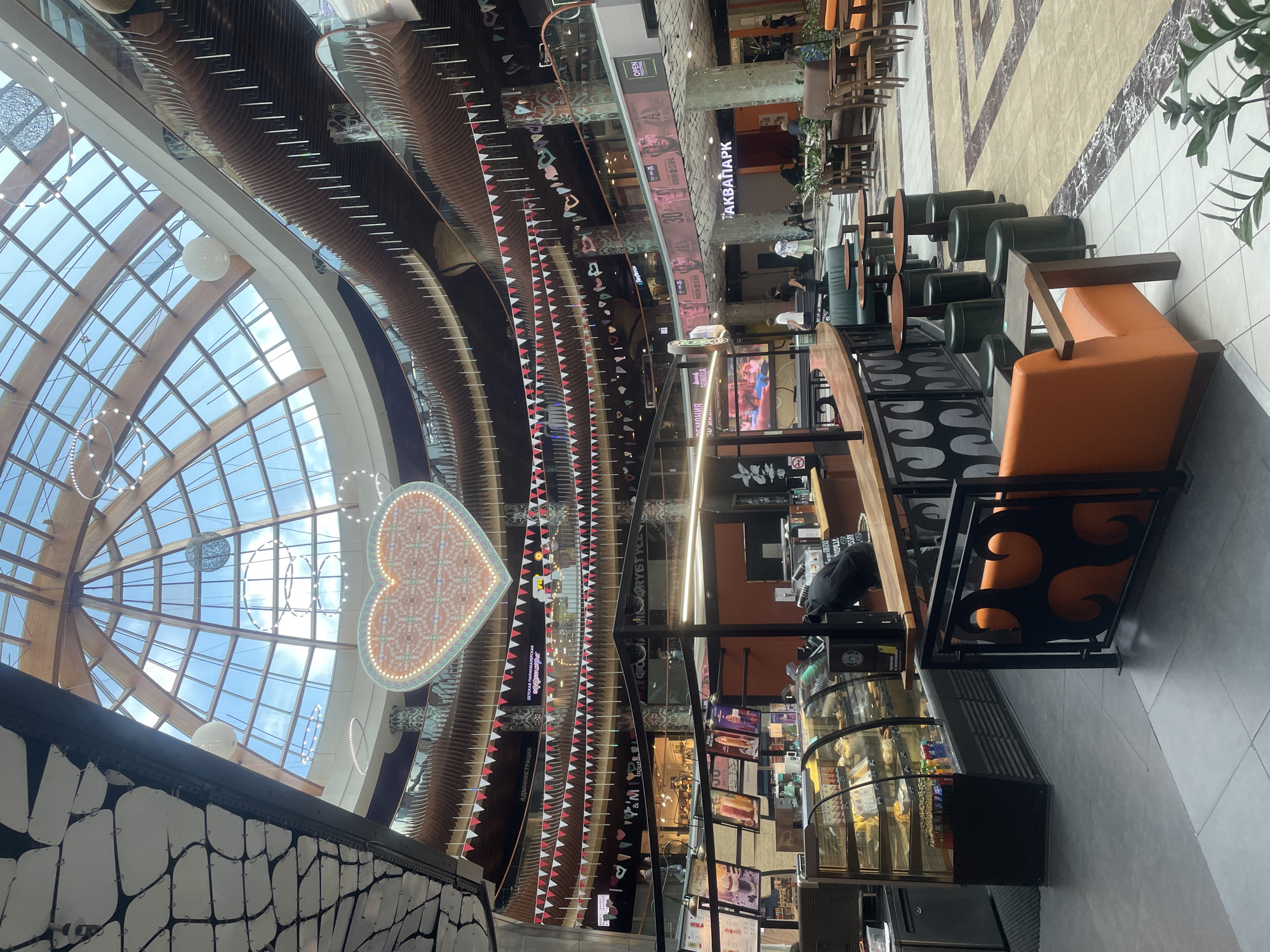 This is an interior shot of the atrium of the Evropa Shopping Center in Saint Petersburg, Russia, on a day with clear skies. The multi-level mall features a large, domed glass ceiling supported by wooden beams, allowing natural light to flood the space. Suspended decorative elements, including string lights and a large heart-shaped illuminated sign, are visible.
The foreground is dominated by a coffee shop counter with a refrigerated display case filled with pastries and drinks. Behind the counter, an employee, dressed in black, is seen preparing orders. Several tables and chairs, some with green stools and others with orange seating, are arranged around the coffee shop.
Further into the mall, a variety of shops are visible on different levels, some with illuminated signage. Banners and strings of small flags are hung across the atrium at various levels. People are seen browsing shops, sitting at tables, and walking through the mall. The flooring is a mix of light-colored tiles with marble-like patterns. The architecture of the mall is characterized by curved wooden accents and railings on the upper levels.