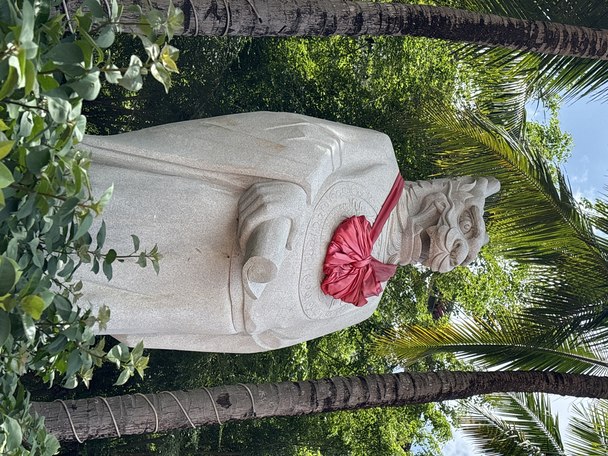 A stone statue of a mythical creature with a dragon-like head stands outdoors, adorned with a large red silk ribbon tied in a bow around its neck. The statue is positioned between two palm trees and amidst lush green foliage. The creature's head is detailed with sharp teeth and an open mouth, and it wears traditional robes. In its right hand, the statue holds a rolled scroll. The background consists of dense green trees and a bright, slightly cloudy sky. The scene appears to be in Xinzheng, China.