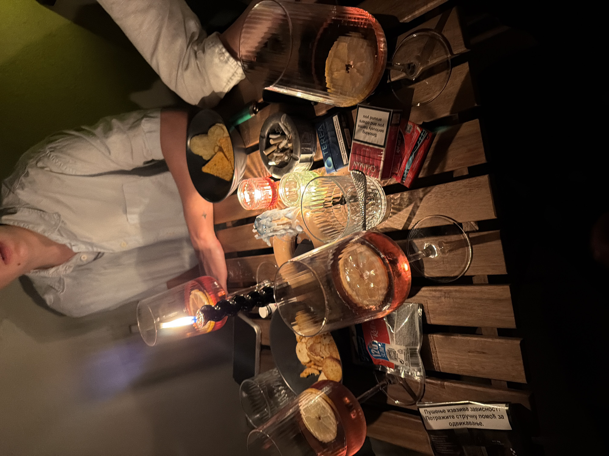 The image captures a convivial gathering at an outdoor location, likely in Palilula Urban Municipality, Serbia, given the visible Serbian text on a cigarette pack warning of addiction. A wooden table serves as the focal point, laden with glasses containing beverages and lemon slices. A person with light skin and wearing a white shirt is partially visible, and their arm rests on the table near a small plate of snacks. The table also holds cigarette packs, an ashtray filled with cigarette butts, and a small, lit candle. A clear, slender object with a blue flame is positioned near the left edge of the table, possibly used for lighting cigarettes. The scene appears dimly lit, contributing to a relaxed and informal atmosphere.
