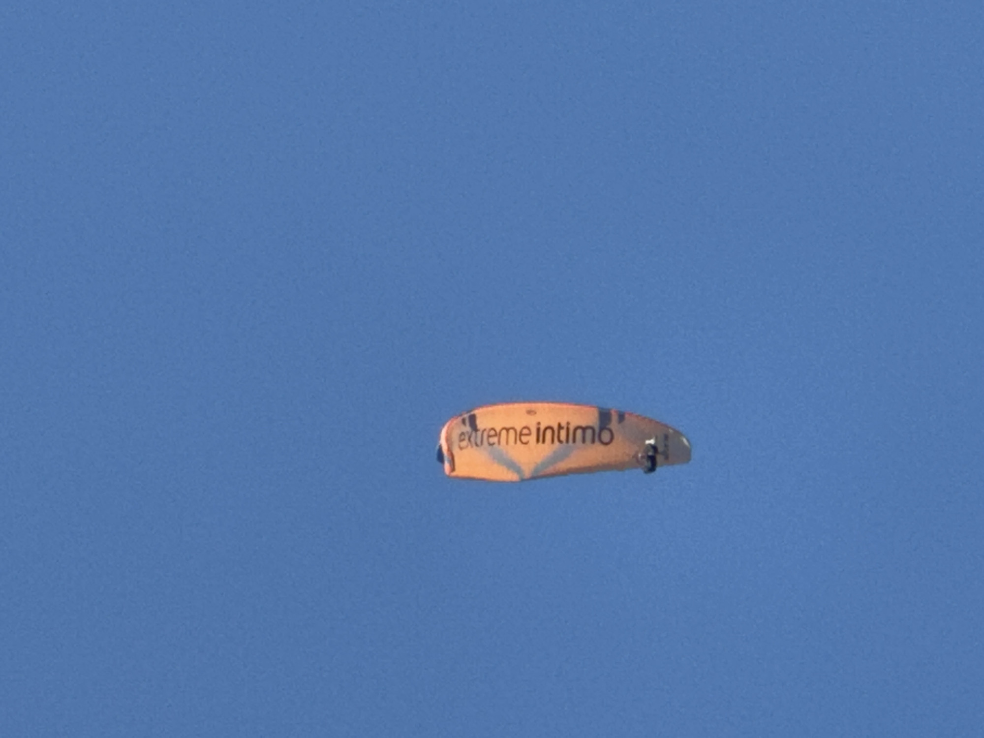A paraglider with orange and yellow markings, reading "extreme intimo," is airborne against a clear blue sky. The paraglider is the sole subject in the image. No specific location in Boreti, Montenegro, can be identified from the provided image.