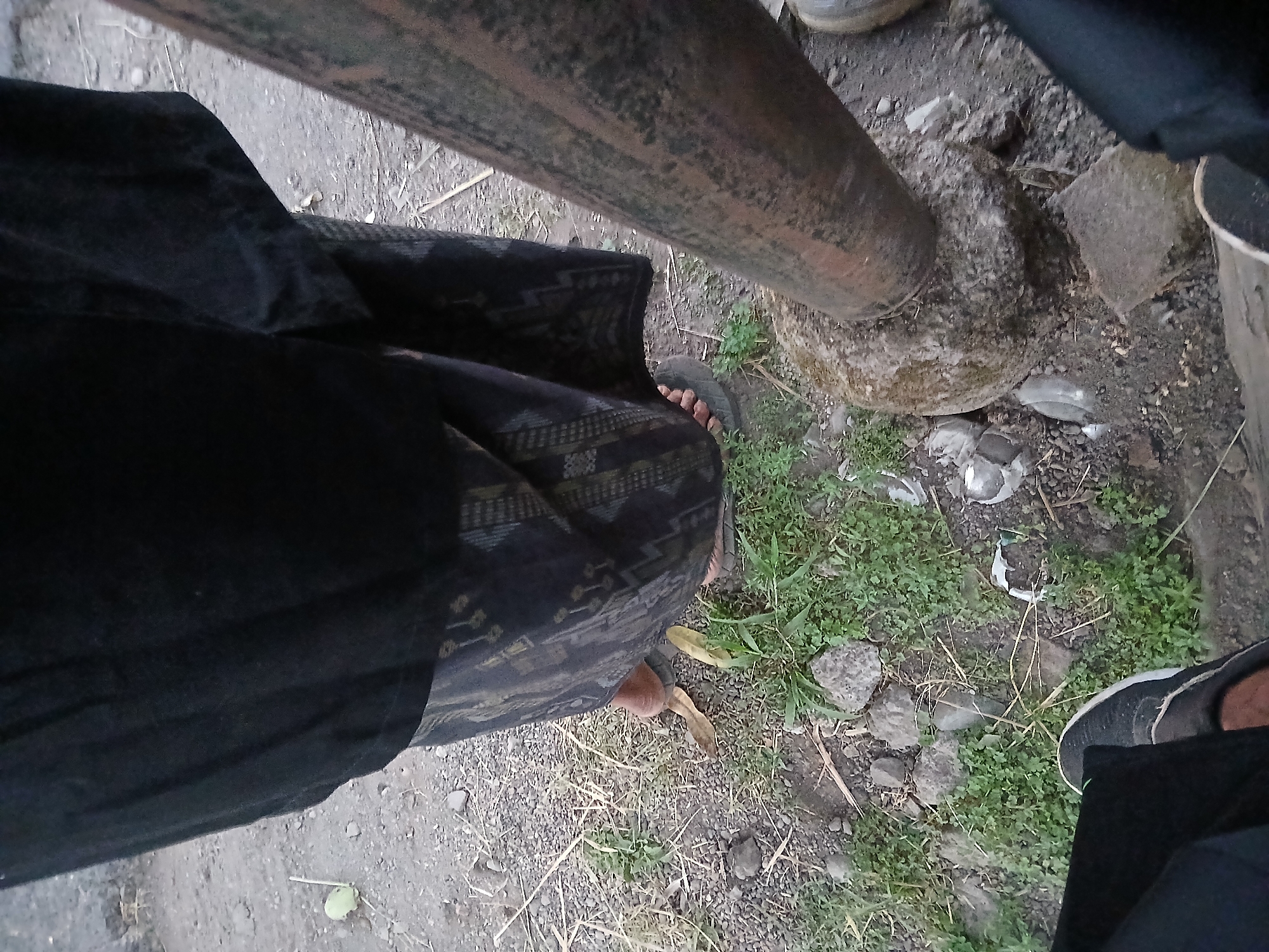 The image shows a close-up, overhead view of two individuals' legs and feet, likely in Kademangan Kulon, Indonesia. The ground is composed of dirt, gravel, and sparse vegetation. In the foreground, a rusty metal pipe or log is diagonally positioned. To the left, a person's legs are visible, wearing dark pants and a dark-colored shoe with a white sole. Further to the right, another person's legs are clad in a patterned dark sarong or wrap. Their feet are bare, with one sandal visible. The overall scene suggests an outdoor setting, possibly a rural or less developed urban area given the natural ground cover and simple attire.
