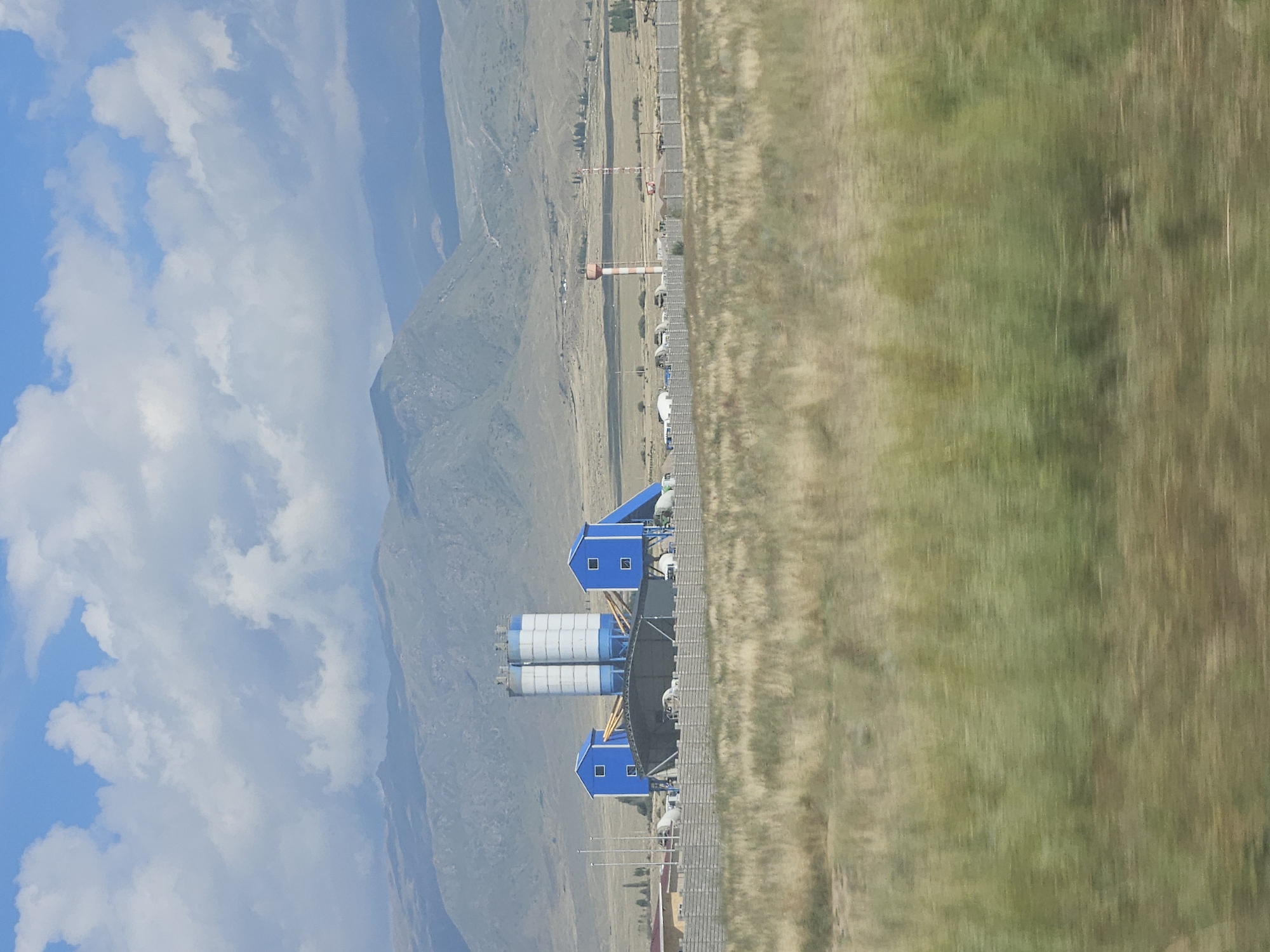 The image depicts an industrial facility situated in a semi-arid landscape with mountainous terrain in the background. The facility features two tall, cylindrical silos painted in white and blue, flanked by two smaller, blue, rectangular buildings with single windows. A large, open-sided structure with a roof covers an area between the smaller buildings. The entire site is enclosed by a concrete barrier fence. In the distance, two tall structures, one resembling a water tower with red and white stripes, and another a striped tower that could be for communication or observation, are visible. The foreground is dominated by dry, grassy vegetation, suggesting a dry climate. The sky above is a mix of blue with scattered white clouds. The overall impression is one of industrial development in a remote, possibly rural, setting.