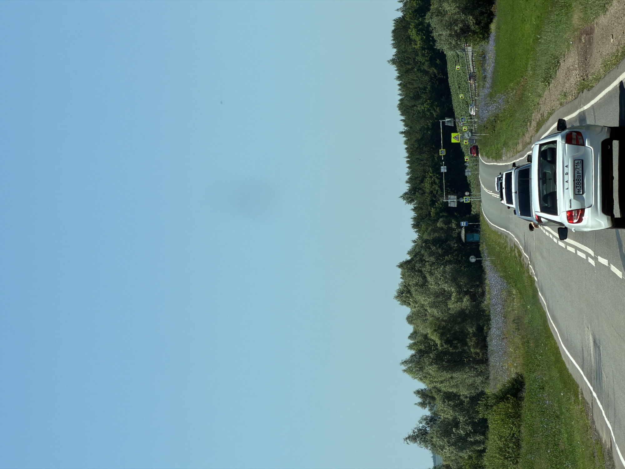 A line of cars is traveling on a paved road through a rural landscape under a clear blue sky. The road has two lanes and white markings. The car in the foreground is white with a license plate that reads "M388BP 716". Other cars are visible further down the road, and some are driving in the opposite direction. The surrounding environment includes green grass, trees, and fields of purple flowers. There are also some traffic signs and a small blue bus stop shelter visible on the side of the road. The scene appears to be a sunny day in the countryside.