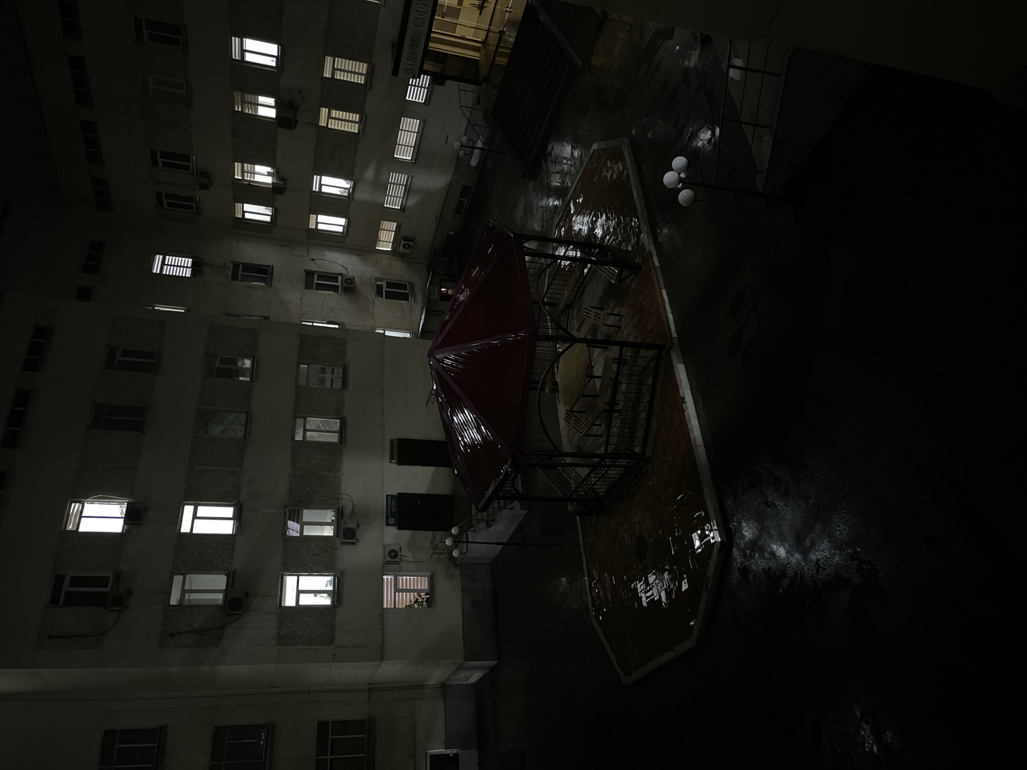 The image, likely taken at night in Navoiy City, Uzbekistan, shows a courtyard surrounded by a multistory building. The courtyard is wet, possibly from recent rain, reflecting light. A red-roofed gazebo sits in the center. Three spherical light fixtures are visible near a shadowed area. The building facade has numerous windows, some illuminated. Air conditioning units are attached to the exterior walls below some windows. The perspective suggests the image was taken from an elevated position.