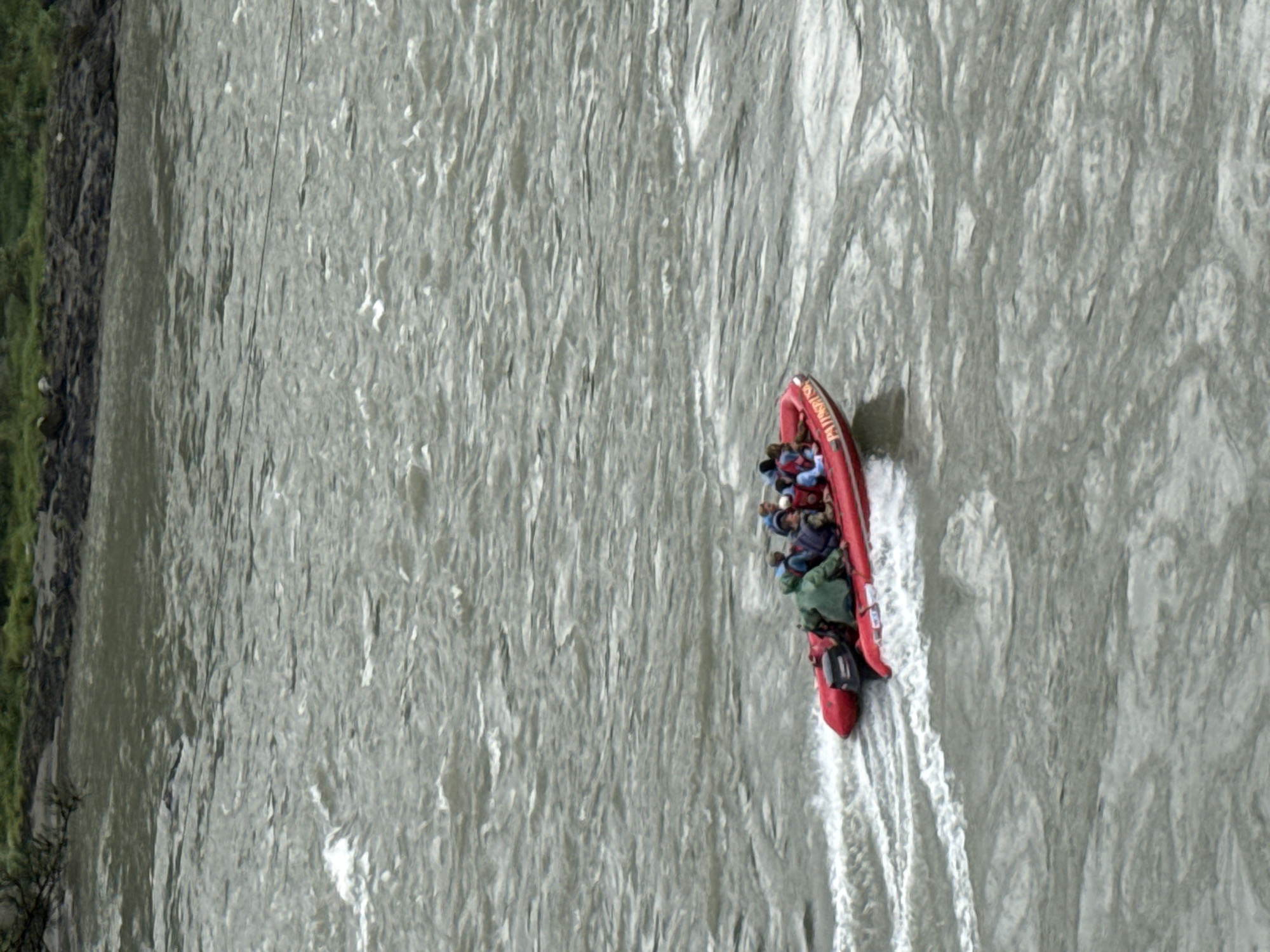 In the city of Barangol, Russia, a red inflatable boat with an outboard motor navigates through choppy, grey water. The boat is occupied by several individuals, most of whom are wearing life vests or jackets in various colors, including green and blue. The water's surface is turbulent, with visible waves and white foam trailing the boat. In the background, the shore is lined with dark rocks and patches of green vegetation. The overall scene depicts a group traveling by boat in a natural, somewhat rough riverine environment.