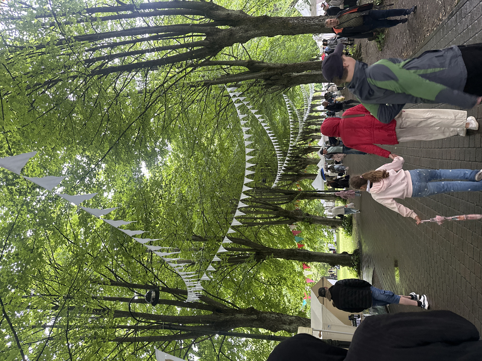 The image depicts an outdoor event, possibly a fair or market, taking place along a tree-lined path in Liepaja, Latvia. The path is paved with brickwork and bordered by large trees with lush green foliage. White triangular flags are strung between the trees, creating a festive overhead canopy. In the foreground, several people are walking along the path. A person in a black hooded jacket is on the far left, partially obscuring the view of a white tent. To their right, a man in a black puffer jacket and blue shorts walks away from the camera. Further down the path, a young girl in a pink hoodie and jeans holds a pink umbrella and walks hand-in-hand with another person wearing a red hooded jacket and cream-colored pants. Several other individuals and tents are visible in the background, suggesting a lively gathering. The lighting suggests it is daytime, with overcast or dappled sunlight filtering through the leaves.