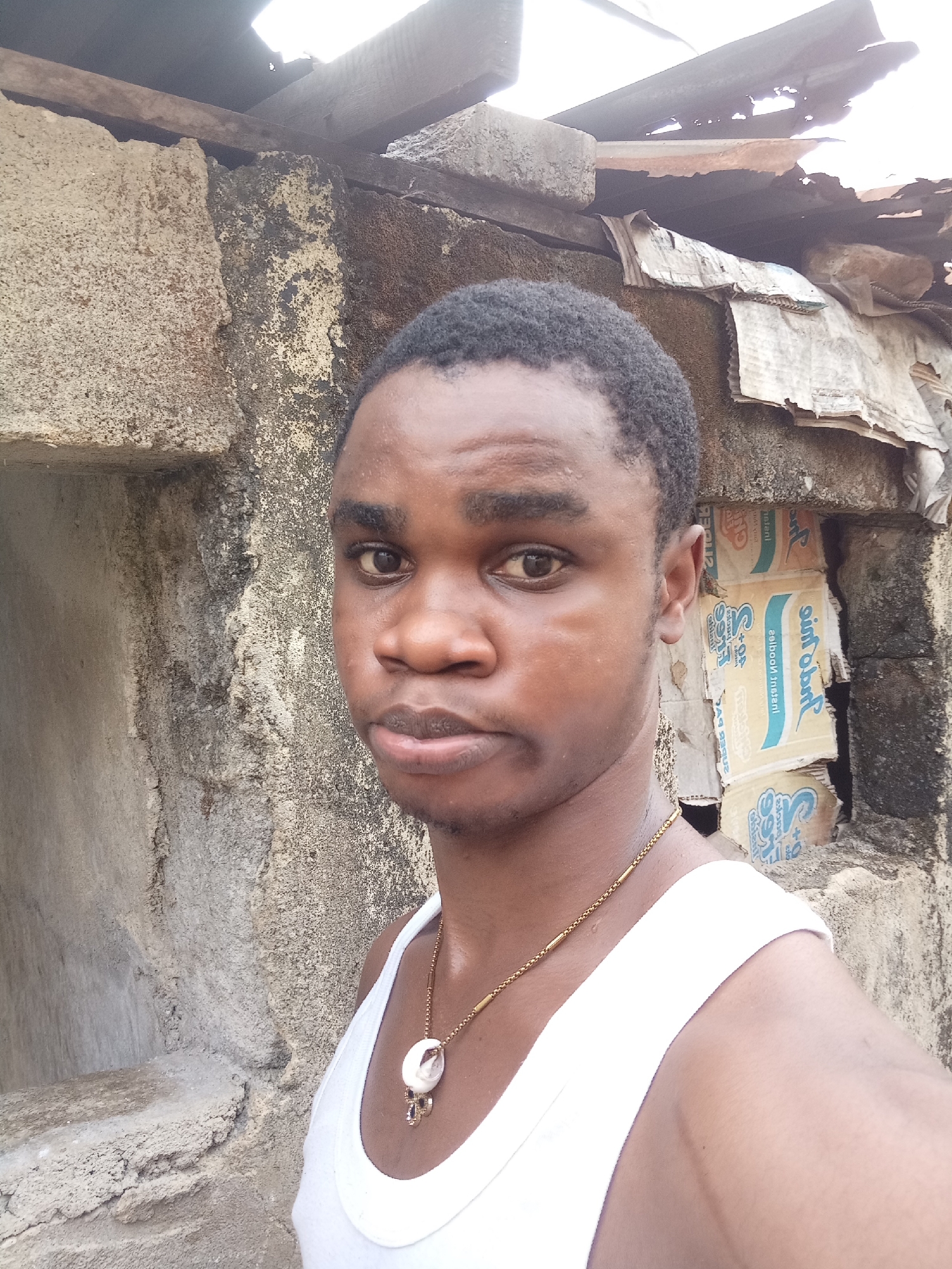 The image depicts a young man with short, dark hair wearing a white tank top and a gold chain with a pendant. He appears to be standing near a weathered, textured wall constructed of rough, light-colored stone blocks on the left. Behind him and to the right is a structure with a makeshift roof constructed from corrugated materials and cardboard boxes. The location seems to be in an urban or semi-urban area based on the architectural style of the construction. The city is specified as Unknown, Unknown.