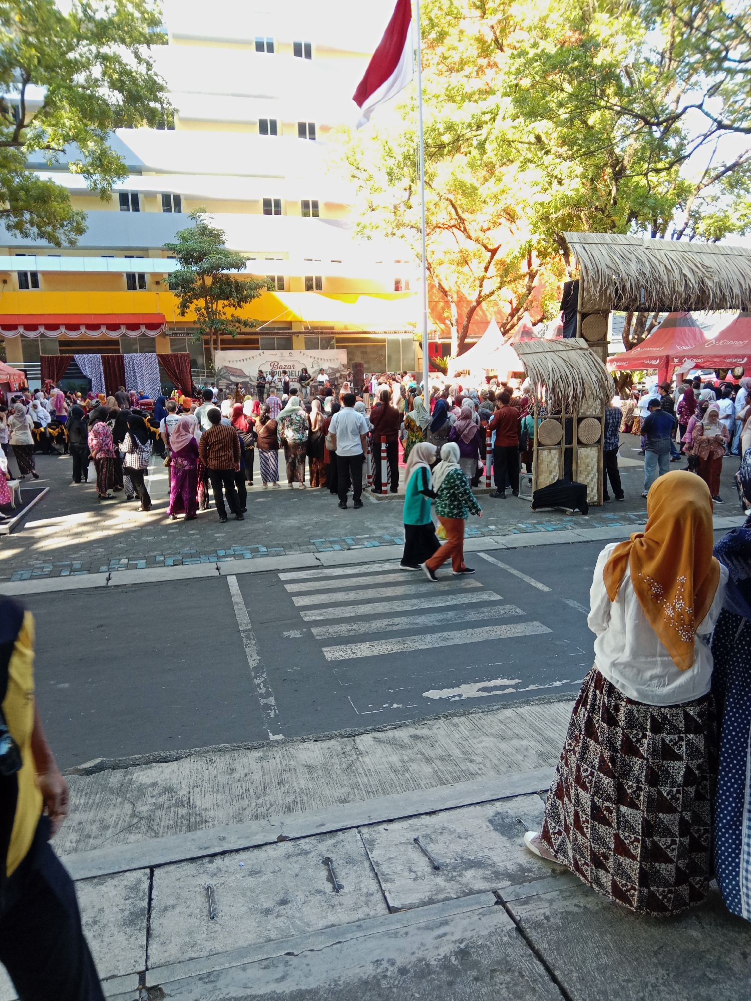 This image depicts an outdoor event, likely a bazaar or festival, in Malang, Indonesia. A pedestrian crossing is visible in the foreground, with several people gathered around. In the background, a modern building with multiple floors and many windows dominates the scene. The building's facade features yellow and white sections. Several tents and a temporary stage with a thatched roof structure are set up, suggesting an organized event. People are seen walking, standing, and observing, many wearing traditional Indonesian attire, including batik patterns and headscarves. A large Indonesian flag is visible flying high above the scene. The environment appears to be a public space, possibly a street or plaza, lined with trees casting dappled sunlight. The overall atmosphere suggests a community gathering or cultural event.