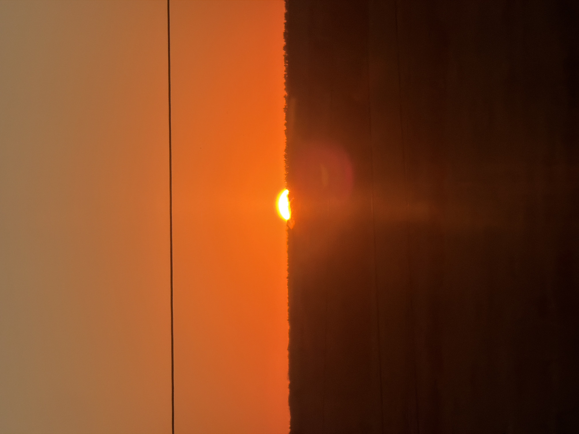 The image depicts a sunset over Karamysh, Russia. A horizontal wire is visible in the upper portion of the frame, bisecting the sky. The sky transitions from a pale orange at the top to a vibrant, fiery orange near the horizon. The sun, a bright semi-circle, is setting just above a dark, silhouetted treeline that stretches across the middle ground. The lower portion of the image is very dark, with the landscape appearing as a deep shadow. A lens flare is present to the right of the sun.