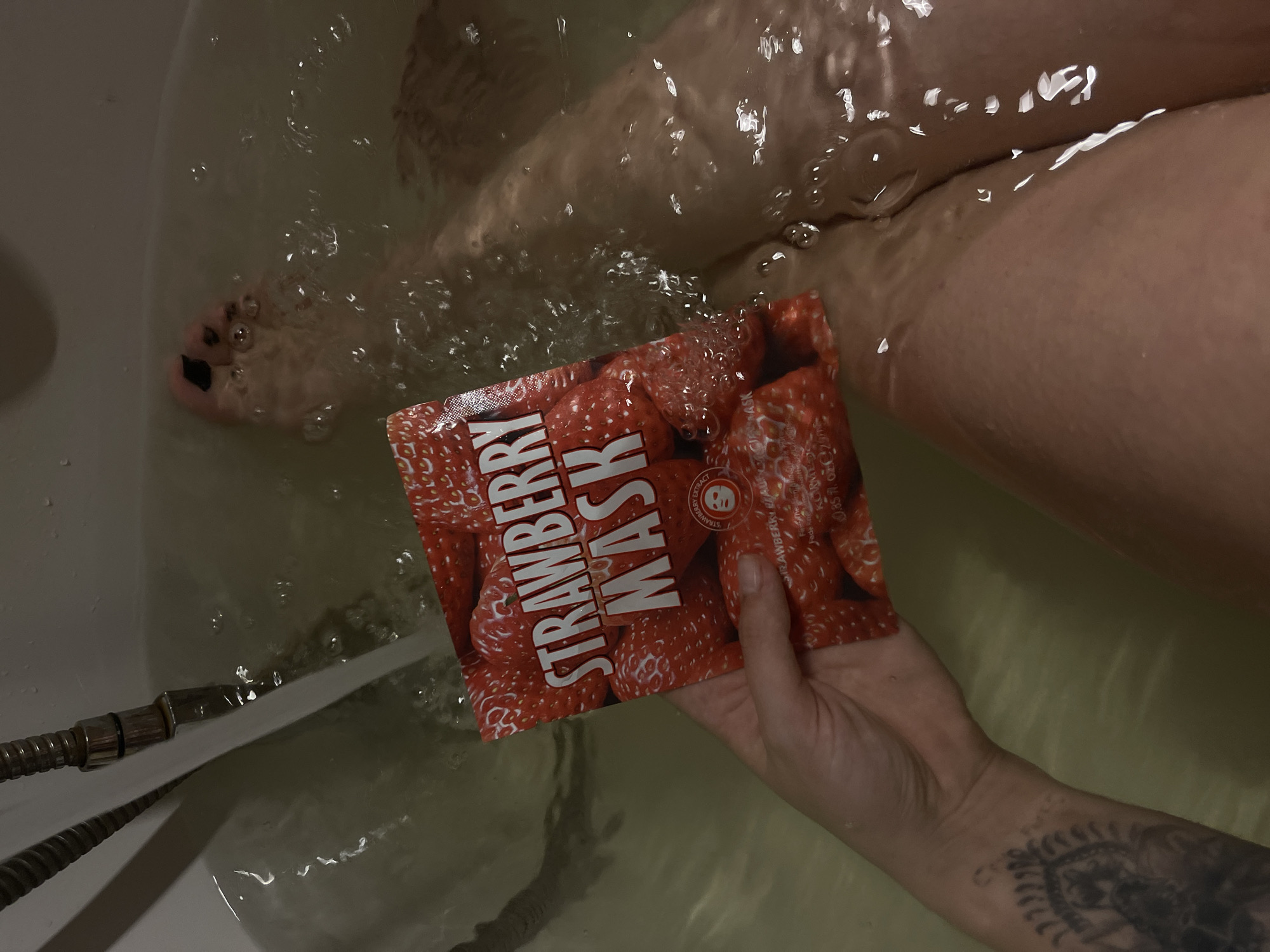 A person soaks in a bathtub. The water is murky. The person holds a strawberry face mask pack. A shower hose and nozzle are present near the edge of the tub, with water flowing into the tub. The person's legs are visible, submerged in the water. Part of the person's tattooed arm is visible. The location in the city of Мосметаллоконструкция Онт, Russia, cannot be ascertained from this image.