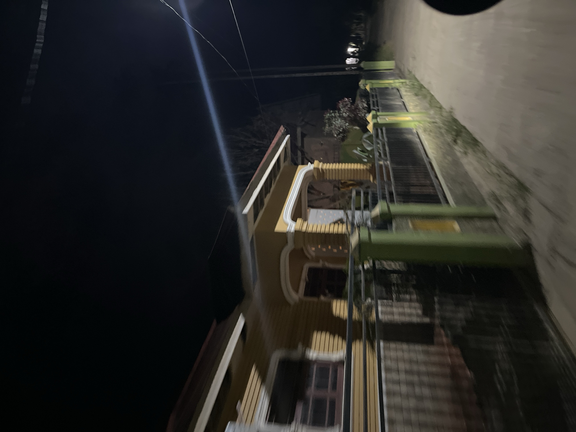 The image shows a nighttime view of a residential street in Menanggini, Indonesia. On the left, a yellow house with white trim is visible behind a fence. The house has several windows with dark interiors, and a covered porch with decorative columns. The fence in front of the house is made of dark metal with green and yellow painted posts. A dirt road runs along the right side of the image, with streetlights visible in the distance. The scene is illuminated by artificial lights, creating long shadows and a slightly blurred effect due to motion. There are no people or animals visible in the image. The environment appears to be rural or suburban, with a dark, clear sky.
