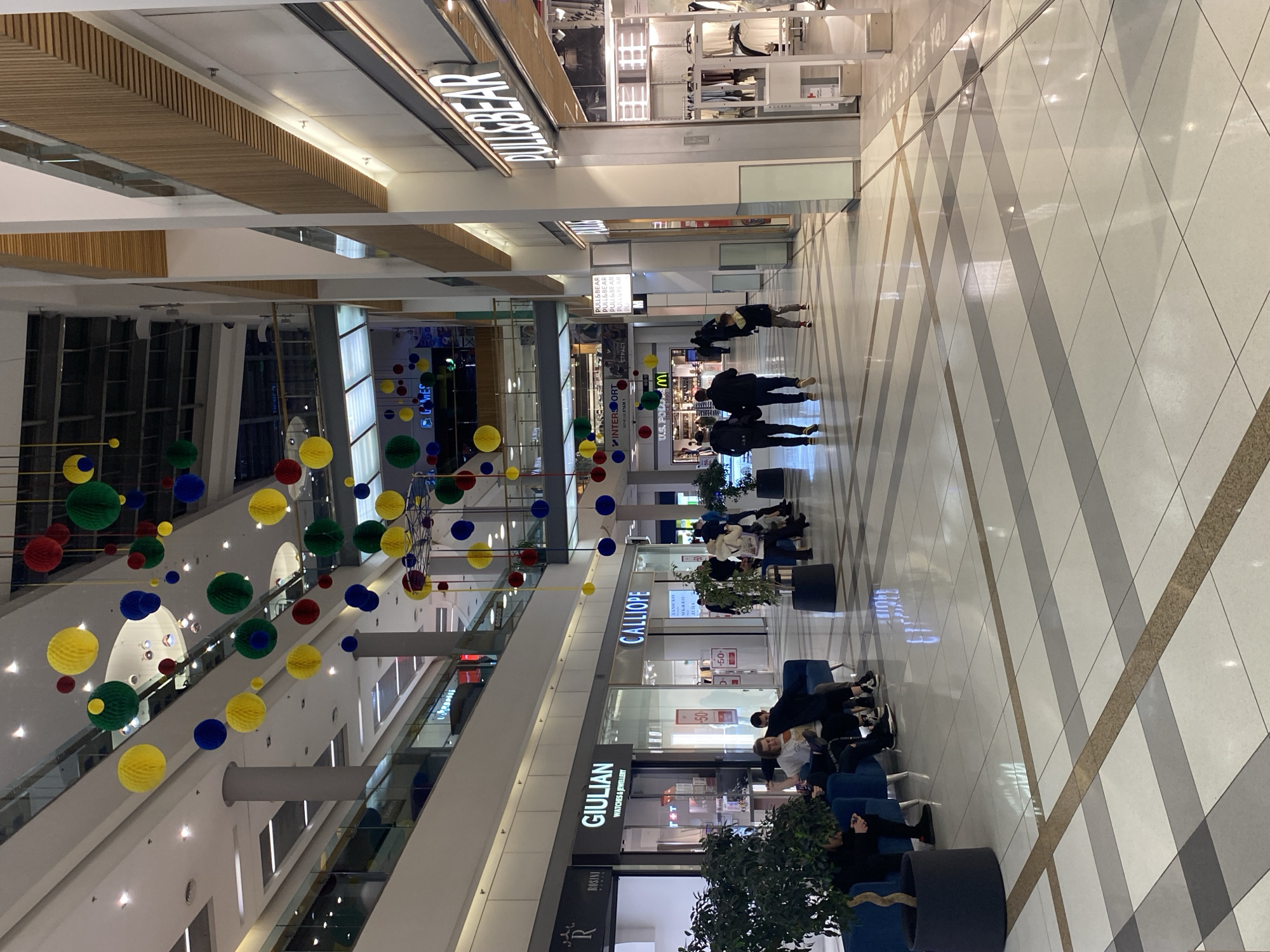 The image shows the interior of a shopping mall. The mall has multiple levels with storefronts visible, including "Giulian Watches & Jewelry," "Calliope," "Intersport," and "Pull&Bear." There are people sitting on blue, cylindrical seats in a common area. The floor has a pattern of diagonal lines. There are decorative hanging spheres of red, yellow, blue, and green colors. The mall appears to be in Varna, Bulgaria, as suggested by the Cyrillic script used in some of the shop signage ("Мъжко" and "ДАМСКО").