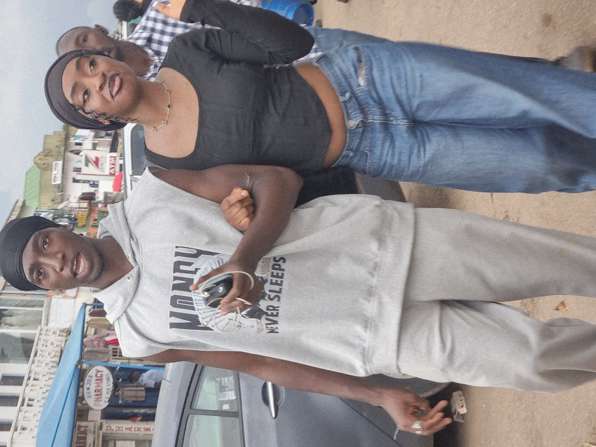 In the city of Jos, Nigeria, a young Black man and woman are captured in a medium shot, walking side-by-side, their arms linked. The man, on the left, wears a grey sleeveless hoodie with the word "MONEY" and the phrase "NEVER SLEEPS" printed on it, paired with grey sweatpants and a black durag. He holds a black object, possibly a phone and keys, in his right hand. The woman, to his right, sports a black long-sleeved crop top and loose-fitting blue jeans, also wearing a black durag and a delicate necklace. Her body is angled slightly away from the camera as she playfully sticks out her tongue. The background reveals a street scene with buildings, including a pharmacy with a blue awning, and other pedestrians. The overall setting suggests a casual, outdoor environment within an urban Nigerian landscape.