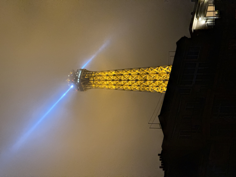 The Eiffel Tower pierces through thick fog, illuminated in yellow light. Two bright blue beams emanate from the top. The tower's latticework structure is visible through the haze. The right side of the frame is dominated by a dark building, only partially visible, with some illuminated windows on the upper floors. The image appears to be taken at night, with a yellow/brown tinge.