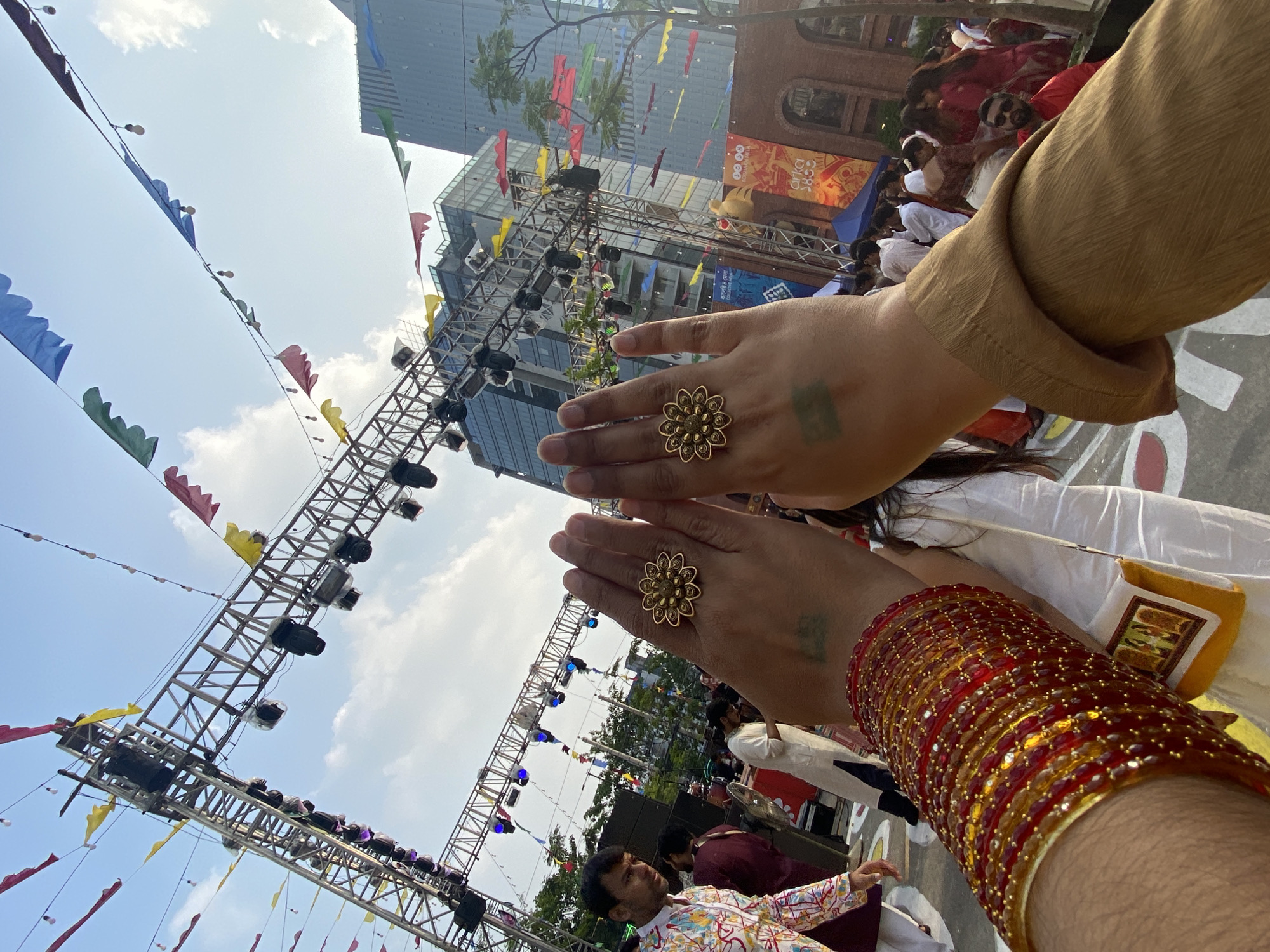 The image depicts a street scene in Dhaka, Bangladesh. Two hands are prominently displayed, one with a golden ring and green ink markings, and the other adorned with red and gold bangles. These hands are positioned in the foreground, partially obscuring the background. The background reveals a festive atmosphere with colorful flags strung overhead and a stage setup with lighting equipment. People are visible in the background, suggesting a public gathering or celebration. Modern buildings are also part of the skyline, confirming the urban setting of Dhaka. The scene appears to be a cultural event or festival taking place in a public space.