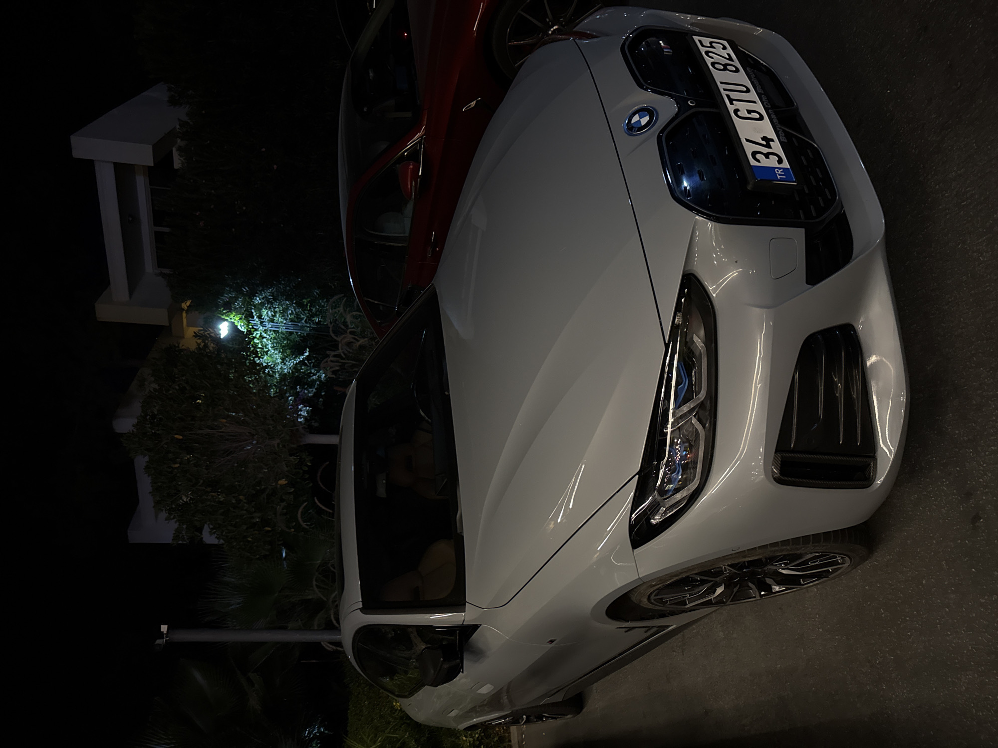 In Bodrum, Turkey, at night, a light gray BMW sedan is parked facing forward in the foreground. The car has a sunroof and its headlights are on, illuminating the road ahead. A red sedan is parked behind the BMW. The background features a modern building with white geometric architectural elements and lush green trees. A lamppost with a bright light illuminates the scene. The license plate of the BMW reads "34 GTU 825".