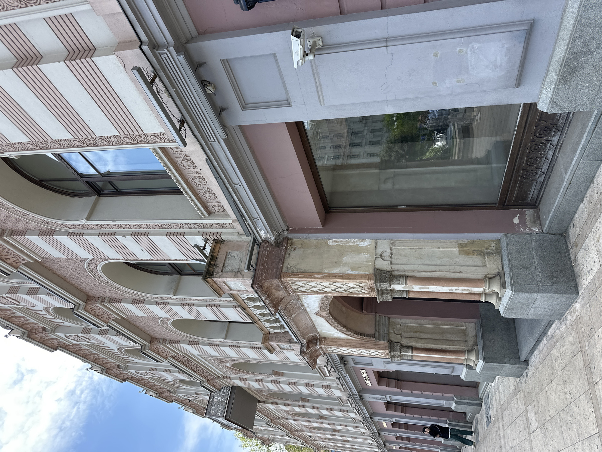The image captures a detailed view of an ornate building facade in Tbilisi, Georgia, oriented as if looking upwards from street level. The architecture features a blend of classical and decorative styles, with light pinkish-beige and white stripes adorning the upper sections and intricate patterned moldings along cornices and window frames.
The building's upper floors display multiple arched windows, reflecting a bright blue sky with scattered white clouds. Below these, there's a large ground-floor window that reflects the surrounding urban environment, including other buildings and green trees. The ground level features sturdy columns with decorative capitals and bases, and sections of the facade are made of light pink and grey stone. A white security camera is mounted prominently above the large window, indicating a focus on surveillance.
On the right side of the image, the grey tiled pavement is visible, where a single individual, a man, is seen standing or walking. He is dressed in dark clothing, including a jacket. The overall impression is one of a historic or well-preserved building under clear daylight conditions. There are no clearly visible or legible texts on any signs or surfaces. The building shows some signs of aging, with minor cracks and discoloration in parts of the stone and plasterwork.
