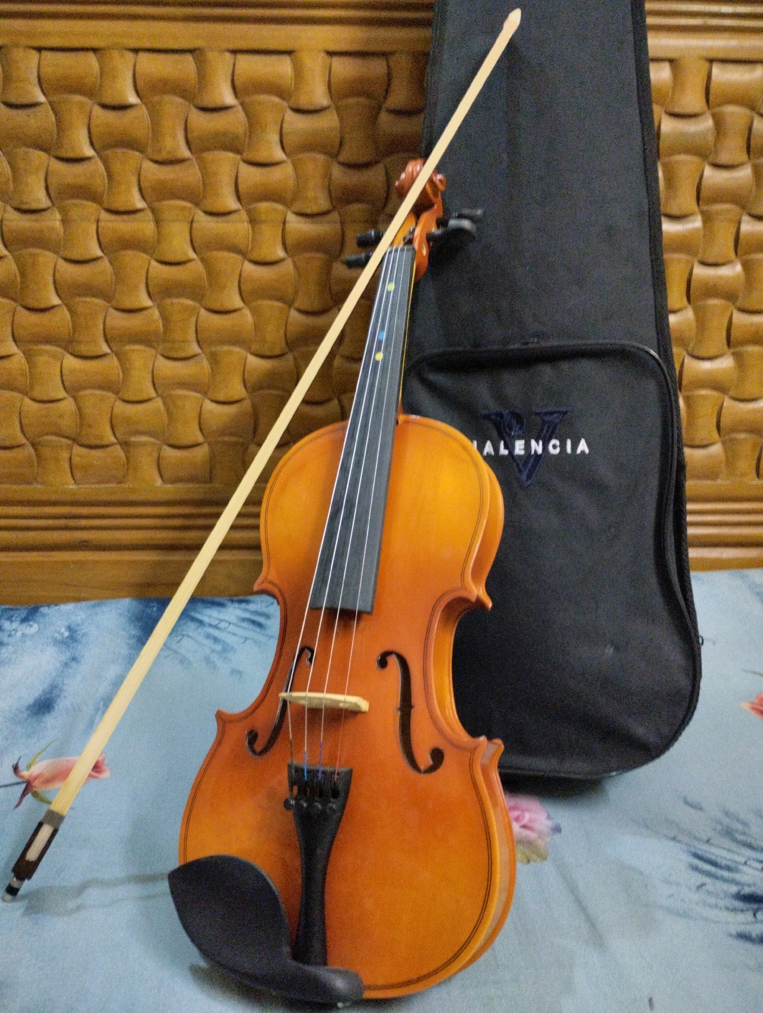 A violin and its bow are positioned in front of a black violin case. The violin is a warm orange-brown color with four strings and a dark fingerboard. The bow is made of light-colored wood with a dark grip. The violin case is plain black with the word "VALENCIA" embroidered in white. The objects are placed on a blue and white patterned surface. In the background, there is a wooden wall with a carved pattern. The scene is lit from the front, casting subtle shadows. The image is a still life composition of musical instruments. No location in Chattogram, Bangladesh is discernible from the image.