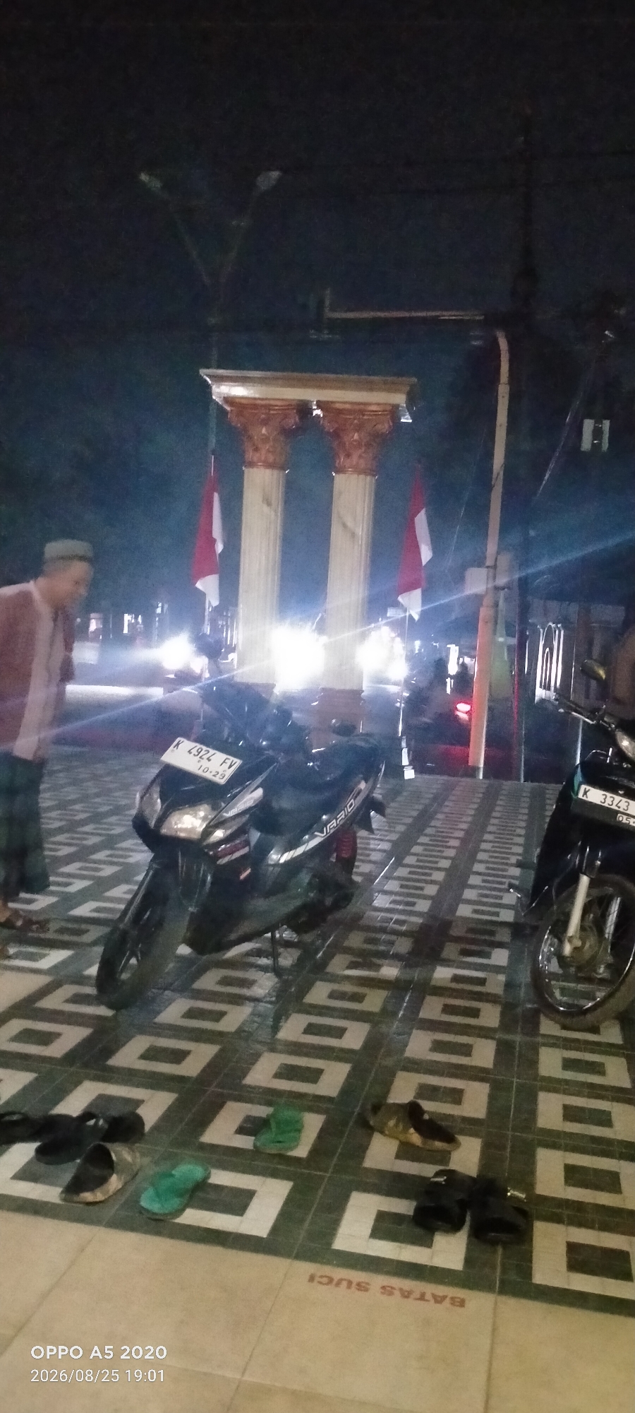 The image captures an outdoor scene at night, likely in the city of Kedung, Indonesia, given the context provided. In the center of the frame, two large, ornate columns stand prominently, topped with a decorative entablature. Two Indonesian flags, red and white, are visible, one on each side of the columns, suggesting a public monument or significant landmark.
In the foreground, two motorcycles are parked. The one on the left is a black scooter with a license plate that reads "K 4924 FV 10-29". The one on the right is also a black motorcycle, with a license plate that is partially obscured but appears to start with "K 3343". The motorcycles are parked on a patterned tiled surface, with a border that reads "BATAS SUCI" in red lettering. Scattered on the tiles are several pairs of footwear, including flip-flops and sandals, indicating that people have likely removed their shoes here.
To the left of the motorcycles, a man is partially visible, wearing a light-colored shirt and a cap. His attire suggests he might be dressed in traditional Indonesian clothing. The lighting in the scene is a mix of artificial lights from street lamps and possibly nearby buildings, creating a somewhat atmospheric, night-time ambiance. The overall impression is a public space at night, possibly near a place of worship or a historical monument, with motorcycles and personal belongings left on the ground.