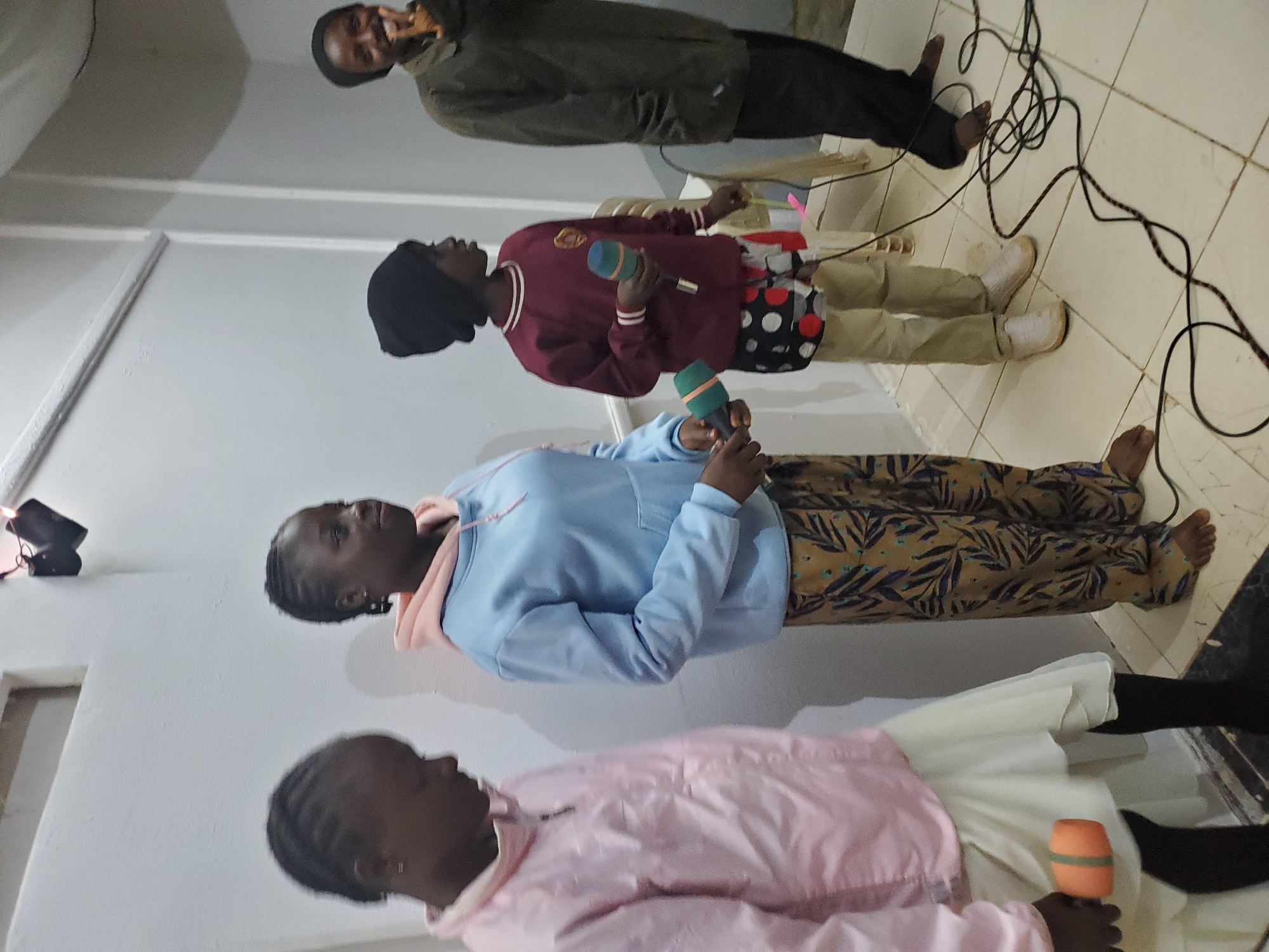 In Kuru Karama, Nigeria, four individuals, three girls and one adult, are positioned in what appears to be an indoor room with white walls and a tiled floor. The room has a ceiling with visible lighting fixtures.
On the far left, a young girl in a pink jacket and a white skirt is holding an orange microphone-like object. To her right, a girl in a light blue hoodie and patterned pants is also holding a microphone. The third individual from the left is a girl wearing a dark beanie, a burgundy sweatshirt, and patterned shorts over tan pants. She is holding a green microphone. On the far right, an adult, partially obscured, is wearing dark clothing and appears to be speaking into a microphone, with their right hand raised and fingers extended.
Cables are visible on the floor, connecting to what might be sound equipment. A chair is also present in the background to the right. The overall lighting suggests an indoor setting, possibly a community hall or a home, with a focus on the group preparing for or engaged in a performance.