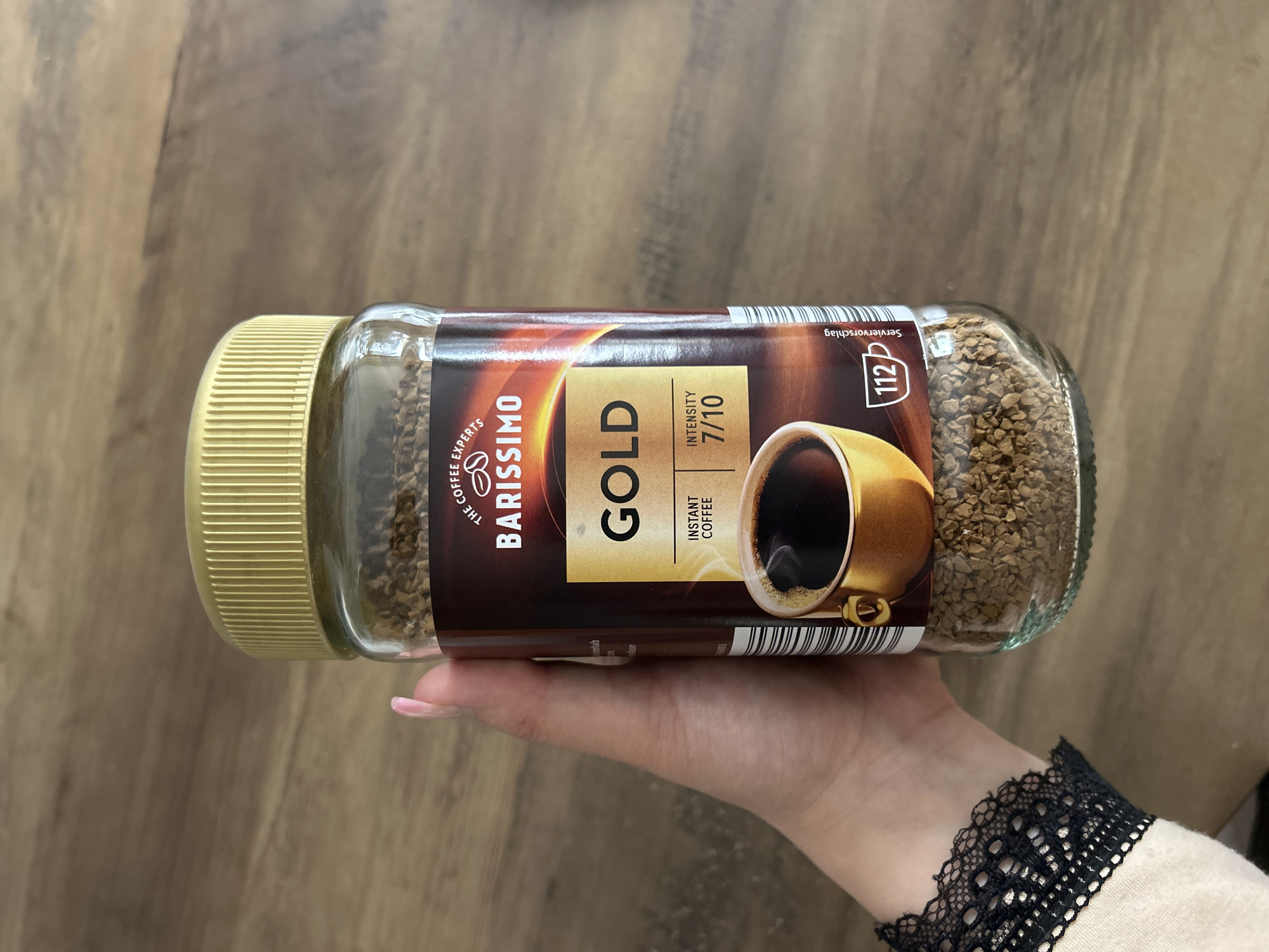 The image shows a close-up of a glass jar of Barissimo Gold instant coffee, held by a person's hand with manicured nails, against a wooden background. The label on the jar prominently displays "BARISSIMO GOLD" and "INSTANT COFFEE" with an intensity rating of "7/10". There is also an illustration of a cup of coffee and the number "112" with a coffee cup symbol. The lid of the jar is gold-colored. The person is wearing a light-colored garment with black lace trim on the sleeve. The wooden surface appears to be a table or countertop. No specific location in Waldbröl, Germany, is depicted or indicated in the image.