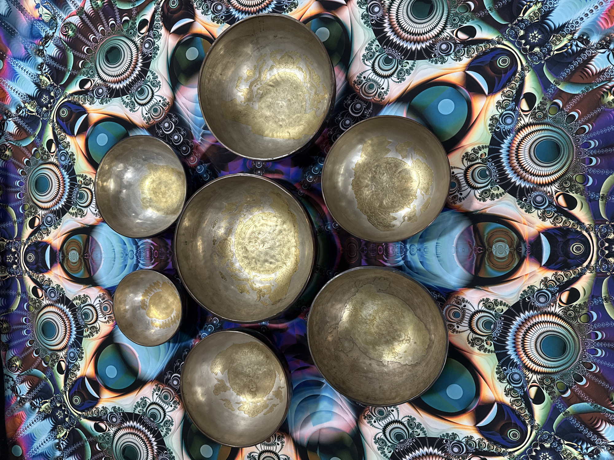 The image shows a top-down view of seven metal bowls arranged in a loose circle on top of a patterned textile. The bowls vary in size and appear to be made of a tarnished silver-colored metal with gold-toned designs or markings inside. The textile has a complex fractal-like pattern featuring repeating circular and eye-like motifs in a variety of colors, including shades of blue, green, brown, and purple. The location is not identifiable as Tashkent, Uzbekistan from the provided image.