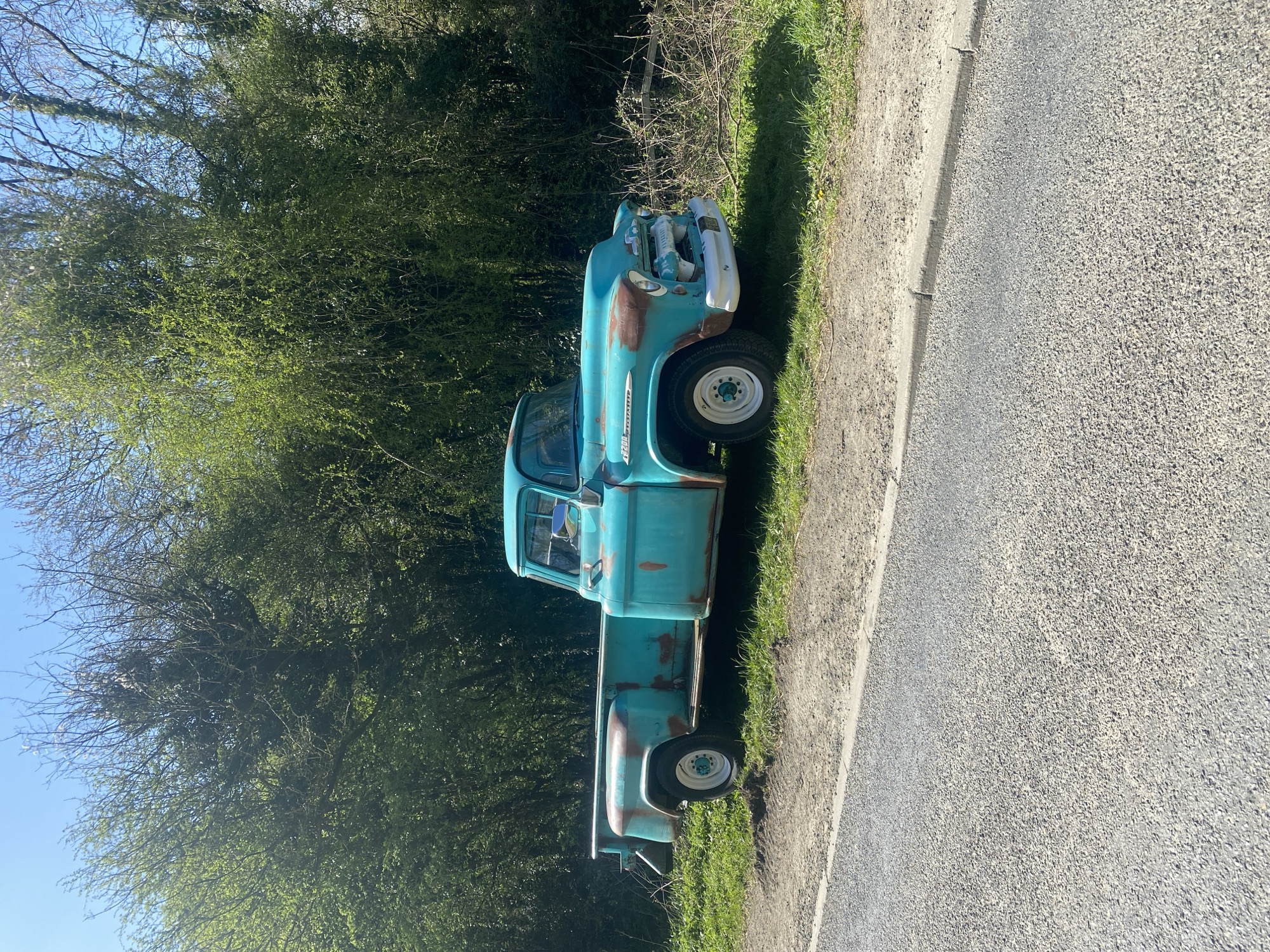 An aged, turquoise pickup truck is parked on the grassy verge adjacent to a road. The truck, exhibiting significant rust, is positioned parallel to the road, its front facing towards the right of the frame. Dense foliage and trees form a backdrop behind the vehicle. The road surface, composed of asphalt, is visible to the right of the truck. The image was taken in Nantmawr, United Kingdom.