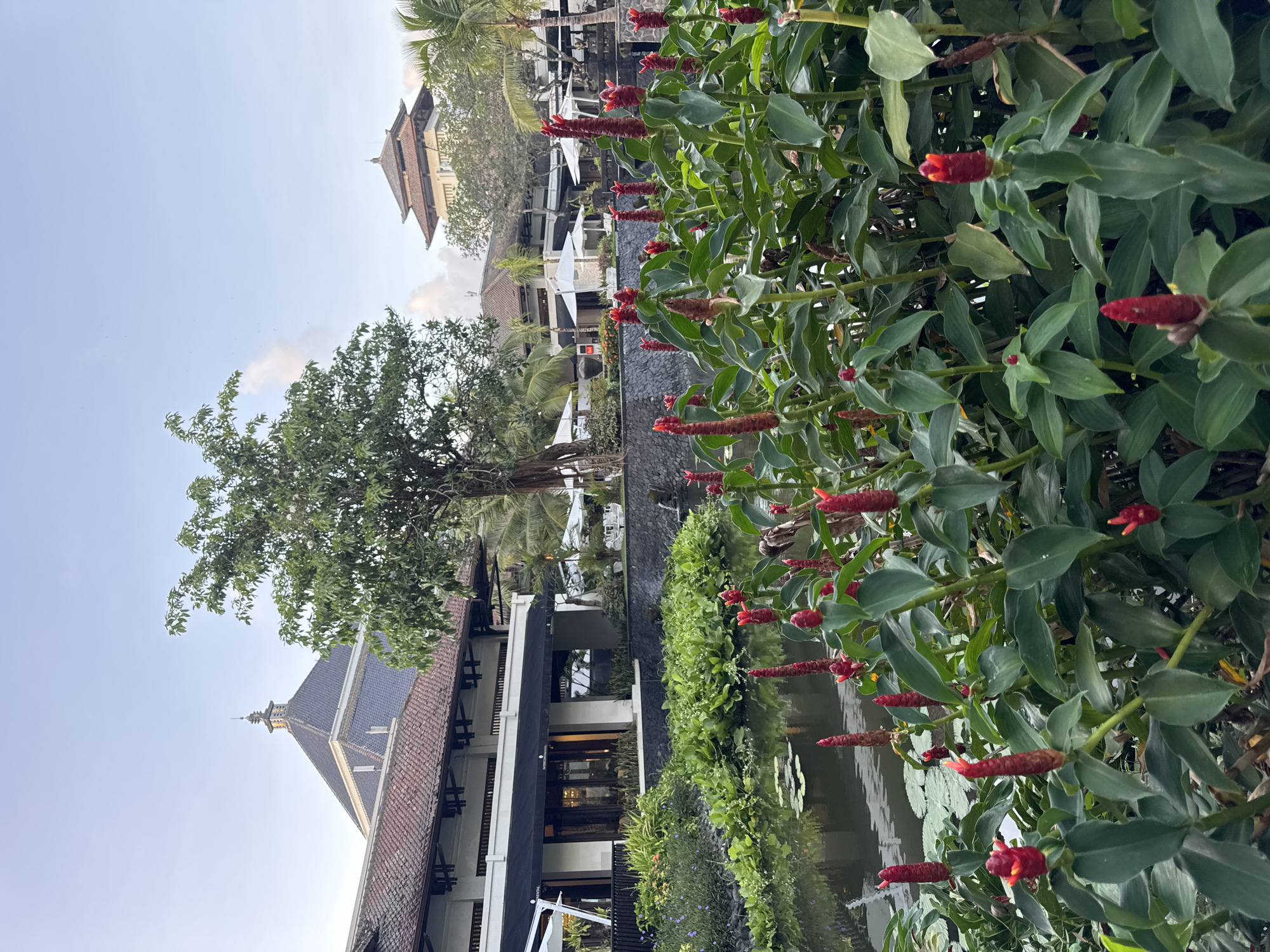 The scene depicts a waterfront area in Kuta Selatan, Indonesia. A building with a dark grey pitched roof and beige walls is visible. In the foreground are lush green plants with red flowers. A large tree stands beside the building. Behind the building, there is a second building with a tiered roof structure. Palm trees are interspersed among the buildings. The sky is light blue with a few clouds. The overall scene appears to be a tranquil, landscaped area. A body of water reflects the surrounding plants and buildings.
