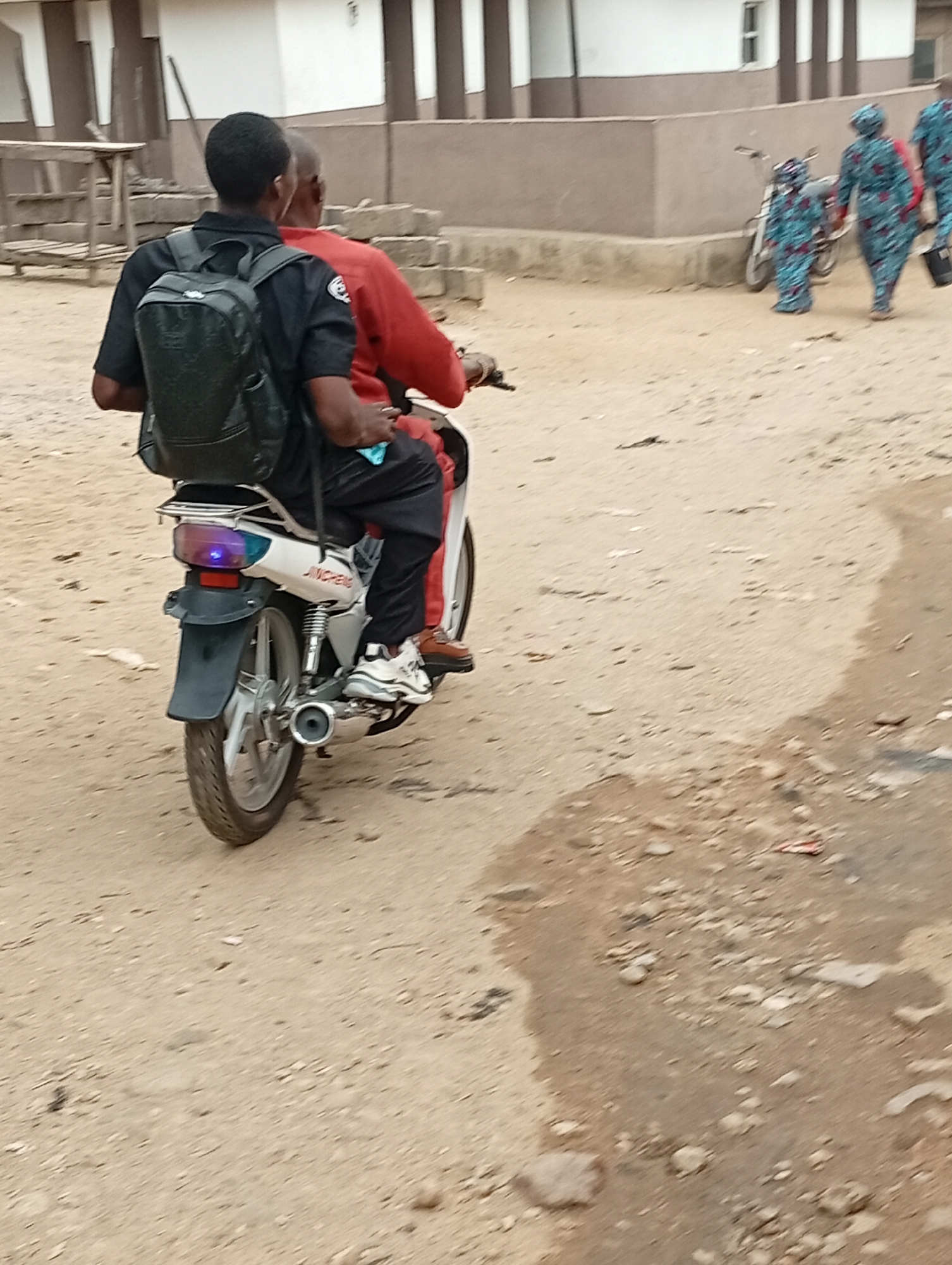 The image shows two people riding a Jincheng motorcycle on a dirt road in Adankolo, Nigeria. The passenger wears a black shirt, black pants, white sneakers, and a black backpack. The driver wears a red shirt, black pants, and brown shoes. In the background, three people in matching blue patterned outfits walk past a parked bicycle. A building is in the background, with white walls and brown trim.