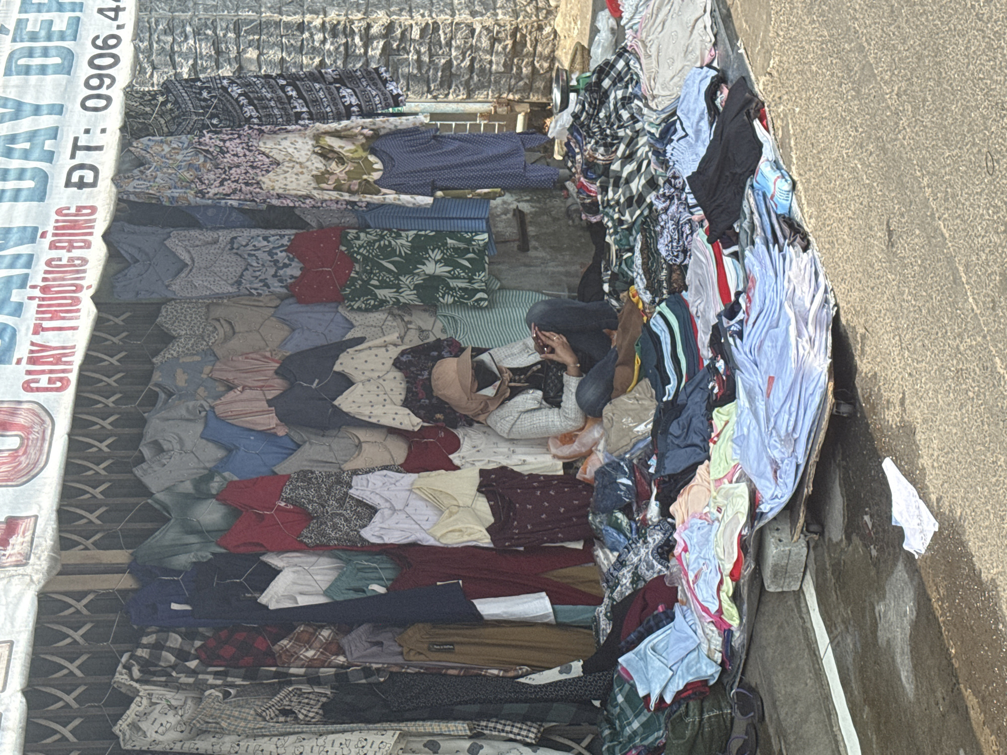 A street-level scene captures a crowded outdoor market in Đà Nẵng, Vietnam. Stacks of used clothing are displayed for sale, covering makeshift tables and ground space. A person is seated amidst the piles, engaging with a mobile phone. The environment is urban, with a building visible on one side and a pavement on the other. A sign with Vietnamese script and phone number "ĐT: 0906.4..." is visible above the clothing. The scene is brightly lit, possibly under direct sunlight.