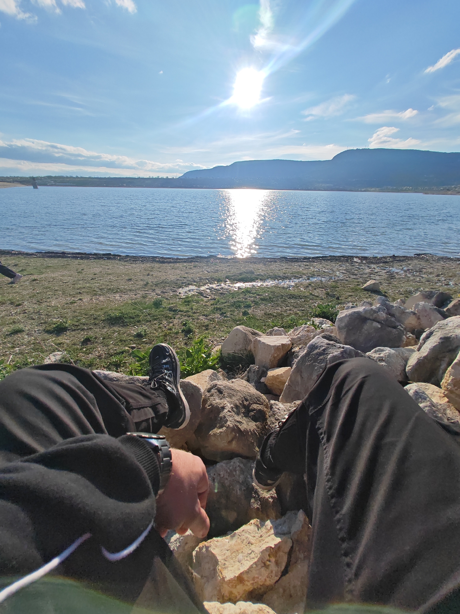 A first-person perspective image shows a person sitting on rocks near a body of water. The person is wearing black pants, a black jacket with a white Nike swoosh, and black and white athletic shoes. The person is also wearing a watch on their left wrist. The body of water is a light blue color and reflects the sun. In the background, there is a mountain range and a blue sky with some clouds. The area near the water is covered with green grass and rocks. The location is near the city of Shumen, Bulgaria, but the precise location within the city isn't provided by the image.
