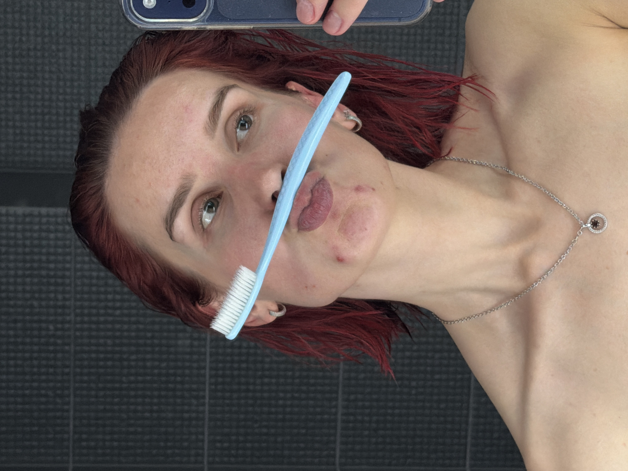 The image displays a close-up, portrait-oriented view of a person's head and upper torso, rotated 90 degrees clockwise. A young individual, likely female, with vibrant red or auburn hair, is looking directly towards the camera with a neutral to slightly playful expression. A light blue toothbrush is held horizontally across their face, obscuring the mouth.
The person wears a silver necklace with a circular pendant, possibly featuring a dark stone, around their neck, and a small silver hoop earring on their visible right ear. In the top right corner of the frame, a portion of a smartphone or camera is visible, held by a hand, indicating that the image is a selfie.
The background consists of a dark gray tiled surface with a distinct grid pattern, suggesting an indoor setting, most likely a bathroom. Minor skin blemishes are noticeable on the person's face. There is no visible text in the image. This casual, personal moment is set in Chaikovsky, Russia.