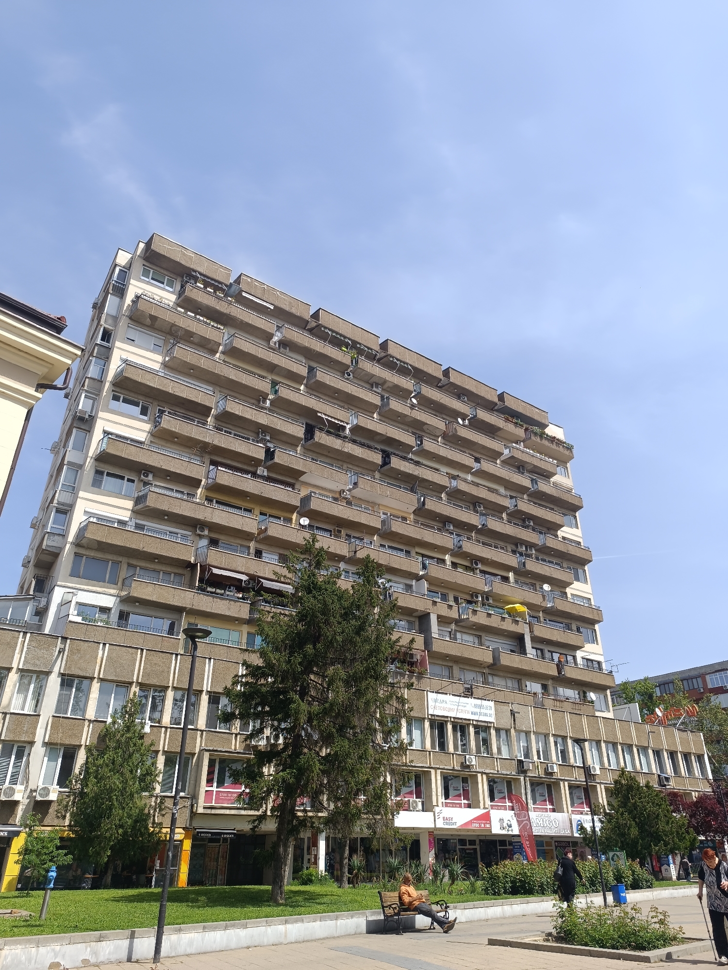 The image captures a tall, multi-story apartment building in Pleven, Bulgaria. The building is characterized by numerous balconies projecting from each floor. Its lower levels appear to house commercial establishments. The street level is bustling with pedestrian activity; individuals are seen walking, while one person sits on a bench. Trees are present, adding greenery to the urban environment. The sky above is clear and blue. The scene conveys a typical day in a city environment.