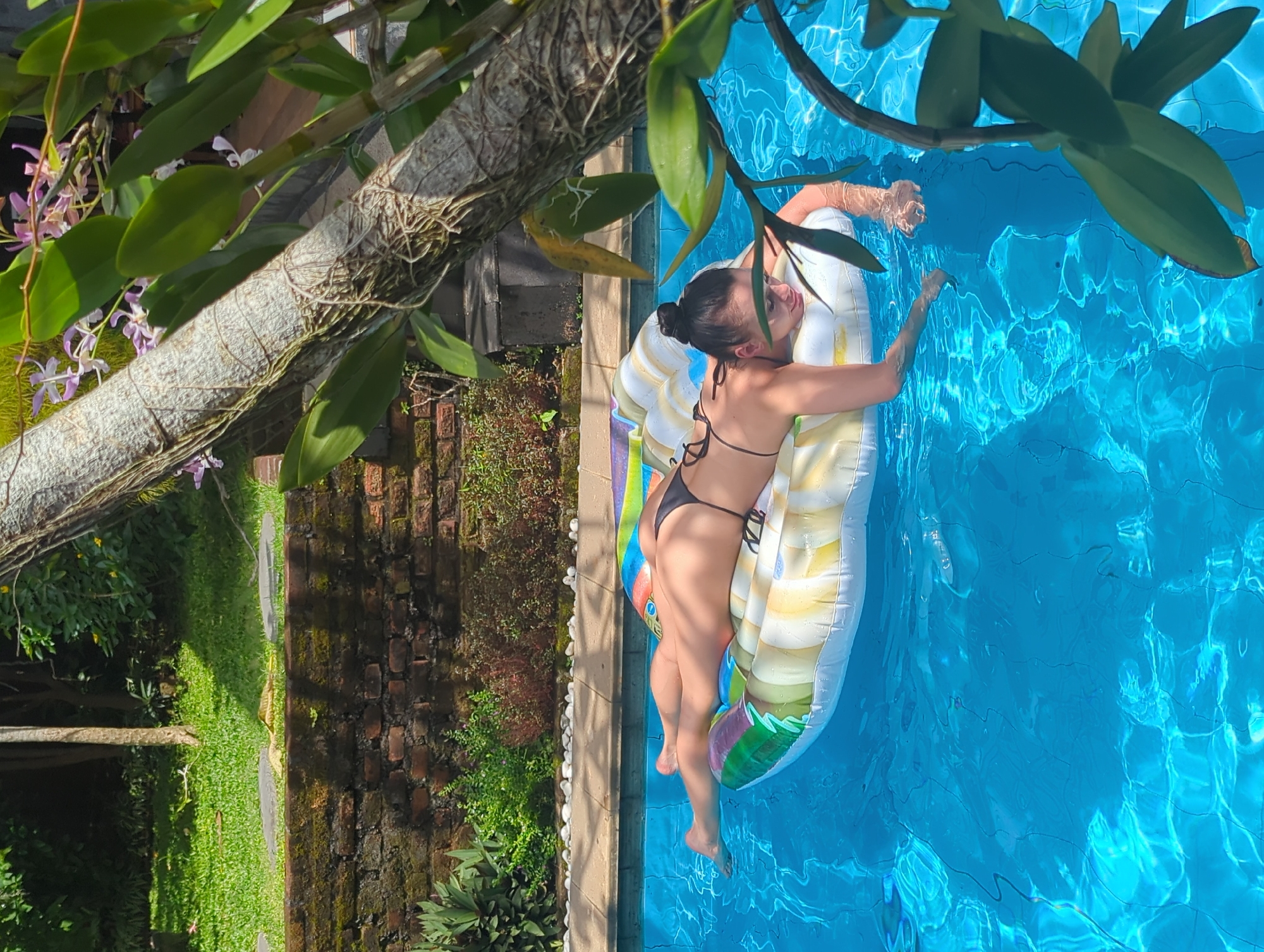 A woman is seen floating on a colorful inflatable raft in a blue-tiled swimming pool. She is wearing a black bikini and has her hair pulled up in a bun. The location appears to be outdoors, likely in North Kuta, Indonesia, given the local vegetation and the architectural style of the surrounding structures, including a brick wall. A tree branch with orchids and green leaves overhangs the pool. The scene is captured from a slightly elevated angle, creating a visually appealing composition that emphasizes both the subject and her surroundings.