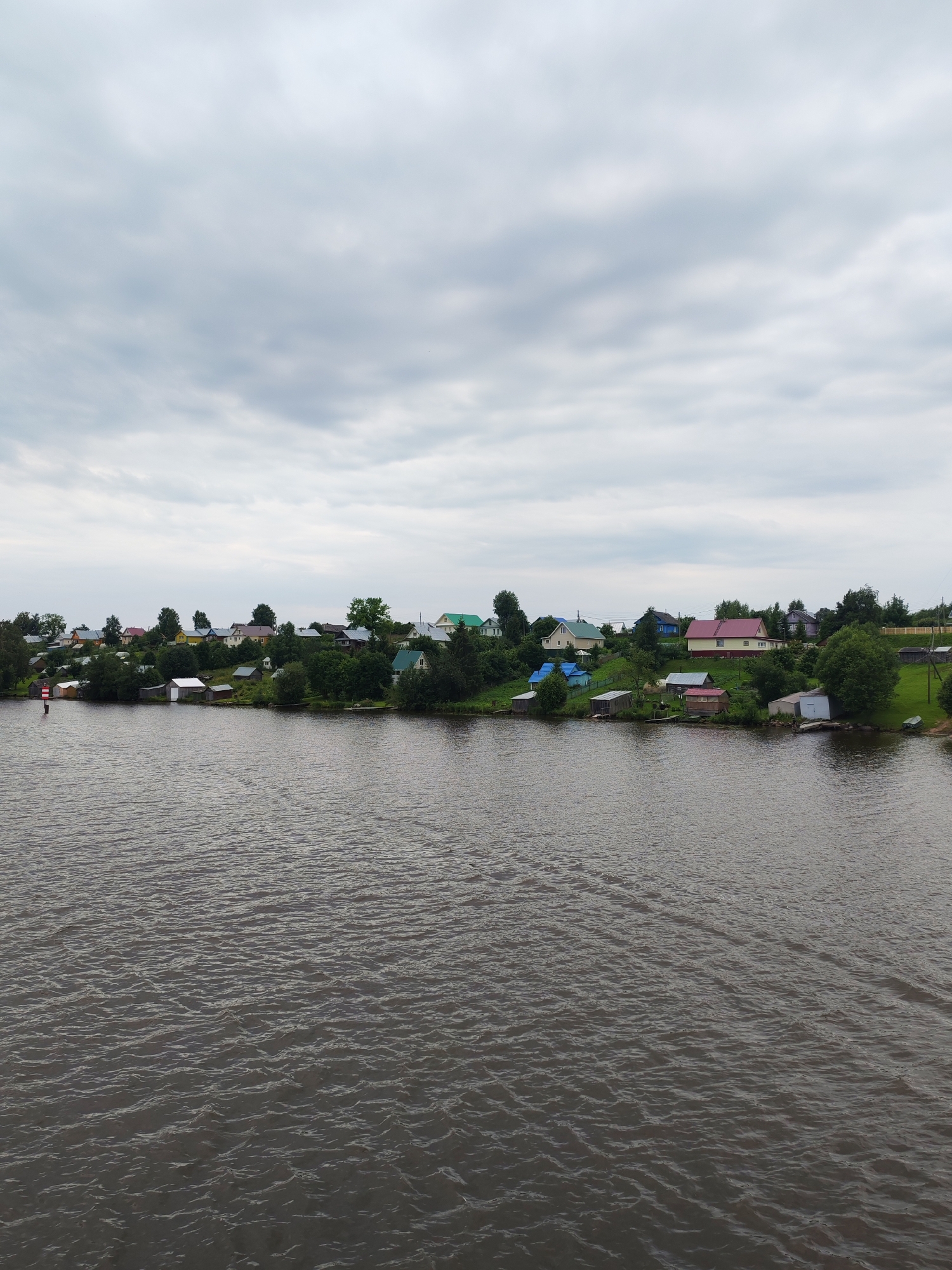 A wide, dark body of water occupies the foreground of the image, its surface rippled and textured. In the background, a verdant, tree-lined bank rises gently, dotted with numerous small, colorful houses. The houses are of various sizes and hues, with roofs in shades of red, pink, blue, and green. Several are situated close to the water's edge, with some appearing to be boathouses or sheds. Lush green trees and bushes surround the structures, creating a sense of a well-established settlement nestled within nature. The sky above is overcast with heavy, gray clouds, suggesting a cool or potentially rainy day. A solitary red and white navigational buoy is visible near the left edge of the frame, marking the water's edge. The overall impression is a serene, rural waterside village scene in Ivanov Bor, Russia.