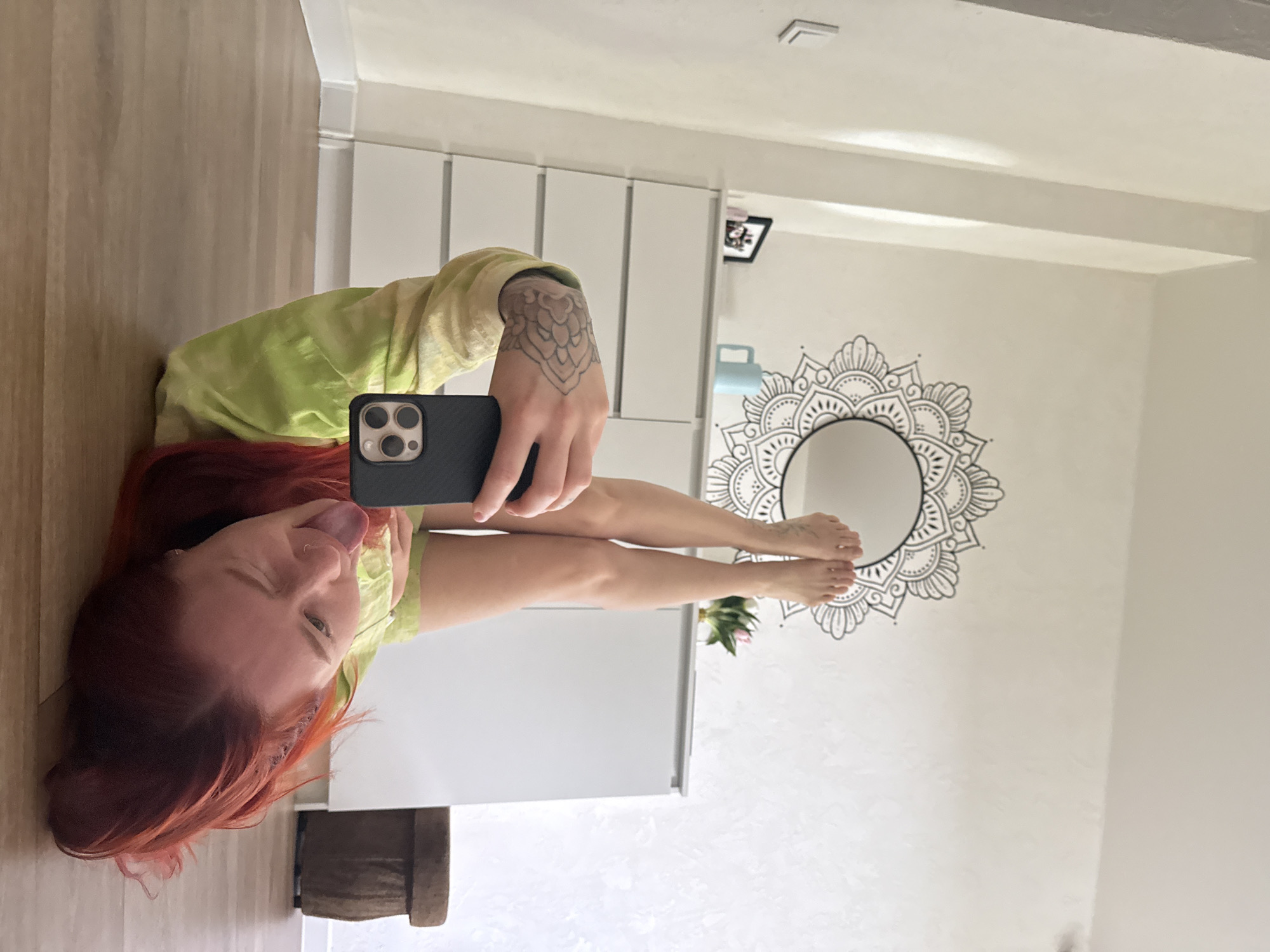 A young woman with bright red hair is lying on a wooden floor, with her legs extended upwards. She is holding a smartphone in her left hand, which is angled to capture a selfie. Her tongue is sticking out, and she is winking at the camera. She is wearing a light green tie-dye t-shirt. In the background, there is a white dresser with several drawers, and above it hangs a circular mandala-style wall decal with a mirror in the center. A small bouquet of pink and white tulips sits on top of the dresser. To the right of the dresser, a round, dark brown stool is visible. The room appears to be a bedroom or living space, with light-colored walls and a light-colored baseboard. The lighting suggests it is daytime, with natural light entering the room from an unseen source. There is no indication of the specific location within Rostov-on-Don, Russia.