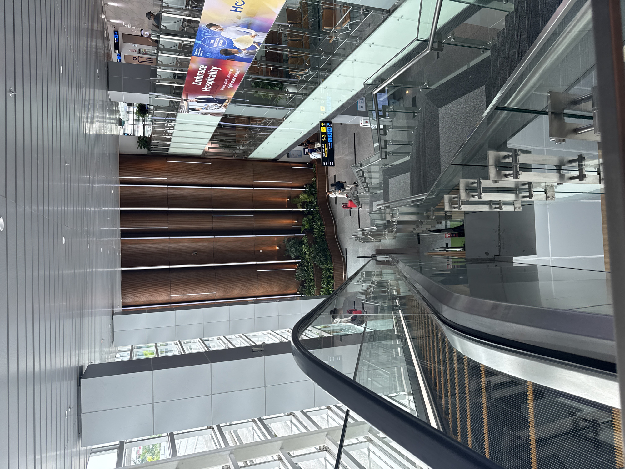 The image shows the interior of the Da Nang International Airport in Da Nang, Vietnam. The perspective is from an elevated position, likely on an escalator or upper level. The scene is dominated by modern architecture, with extensive use of glass, metal, and wooden paneling.
Key elements include:
* **Escalator:** An escalator is prominently featured in the foreground, with a glass balustrade.
* **Airport Interior:** The airport's interior features a combination of textures and materials, including metal ceilings, wood paneling along the wall with recessed lighting, and gray flooring.
* **Signage:** There is a sign with arrows and numbers, indicating directions to different gates (1-3) and other zones. The sign is written in Vietnamese and English.
* **Advertisement:** A large advertisement, "Embrace Hospitality," promotes the airport's amenities, highlighting a focus on taste and care.
* **People:** A few individuals are visible on the level below, including what appears to be a couple walking.
* **Greenery:** There are plants arranged along the wooden paneling.
* **Staircase:** There is a visible staircase with transparent glass and metal supports in the mid-ground.
* **Lighting:** The scene is well-lit, with a combination of natural light from the windows and artificial lighting from the recessed lights.