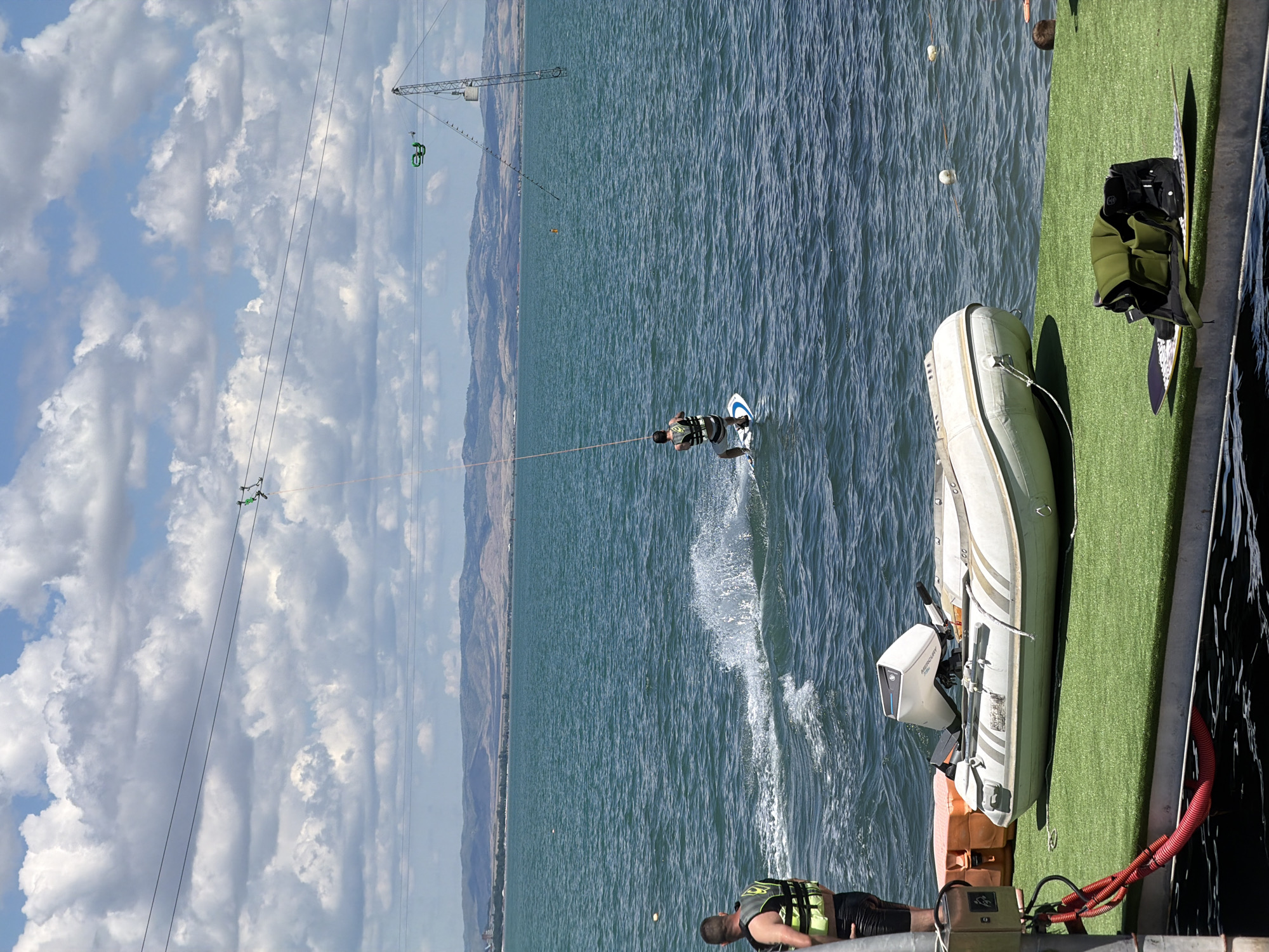 The image captures a scene at the Wake Park on the northern beach of Burgas, Bulgaria. In the foreground, a white inflatable boat with a Mercury outboard motor is docked on a green platform adjacent to the sea. A person wearing a life vest and shorts stands on the left, facing away from the camera. Further out on the turquoise water, another person is wakeboarding, creating a splash behind them, pulled by a cable system overhead. In the background, a distant, hazy shoreline with hills and buildings stretches across the horizon under a partly cloudy sky. The wake park's cable system, with its towers and wires, is visible in the mid-ground.