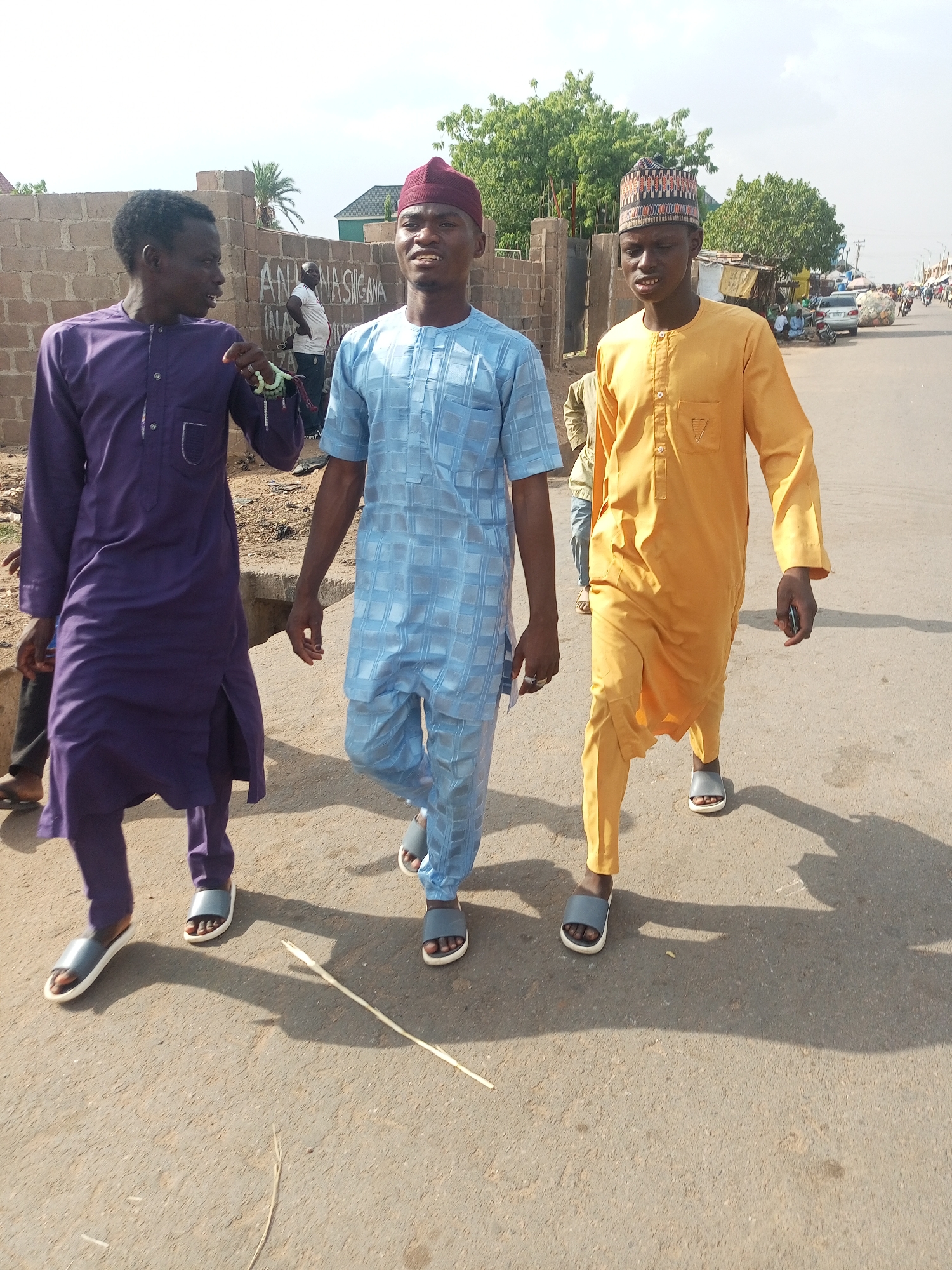 Three young men in traditional attire walk on a paved road in Unknown, Unknown. The man on the left wears a purple robe, the one in the middle wears a blue checkered robe, and the man on the right wears a yellow robe with a matching hat. All three wear similar sandals. In the background is a building with graffiti. Other buildings, vehicles, and people are visible in the distance. A long, thin stick lies on the road.
