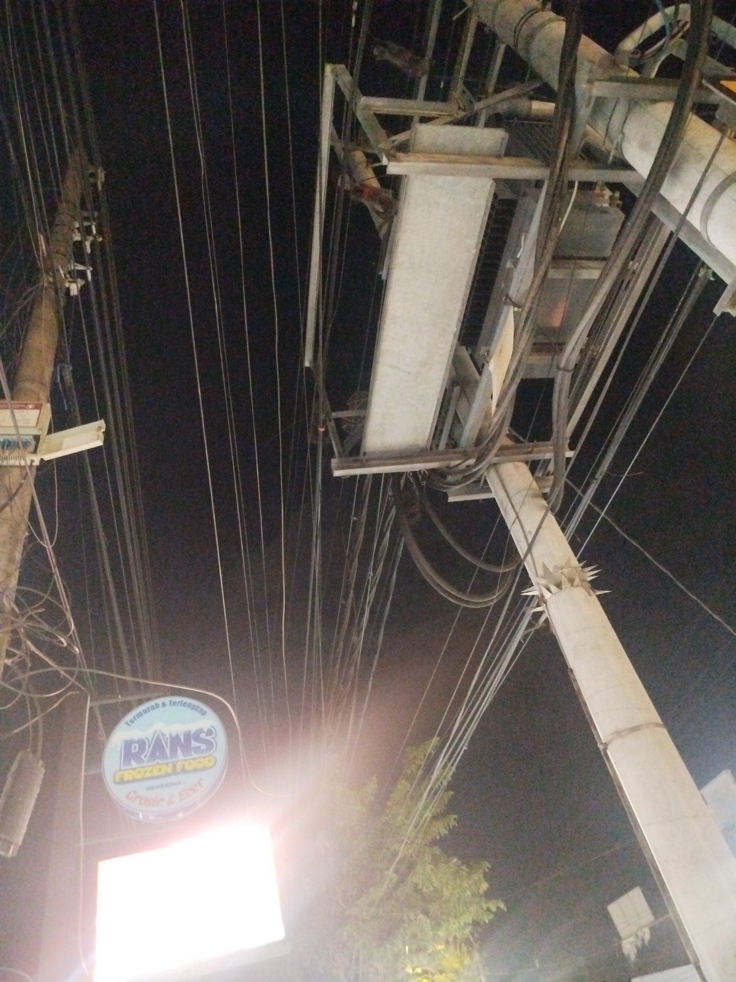 The image captures a nighttime view of a street scene in Burneh, Indonesia, dominated by a dense network of overhead electrical wires. In the foreground, a brightly lit oval sign for "RANS FROZEN FOOD" hangs, displaying text in Indonesian, including "Termurah & Terlengkap" (Cheapest & Most Complete) and "Grosir & Ecer" (Wholesale & Retail). Below this sign is a large, illuminated rectangular sign, possibly for advertising or direction, emitting a strong white light that causes lens flare.
The scene is characterized by a concrete utility pole on the right, from which numerous wires extend upwards and across the frame. A complex assembly of metal structures, likely supporting the electrical infrastructure, is visible above the RANS sign and extending towards the top of the image. The background is dark, indicating it is nighttime, with some foliage from a tree faintly visible to the left, illuminated by artificial light sources. The overall impression is one of urban infrastructure with commercial signage, typical of a streetscape in Indonesia at night.