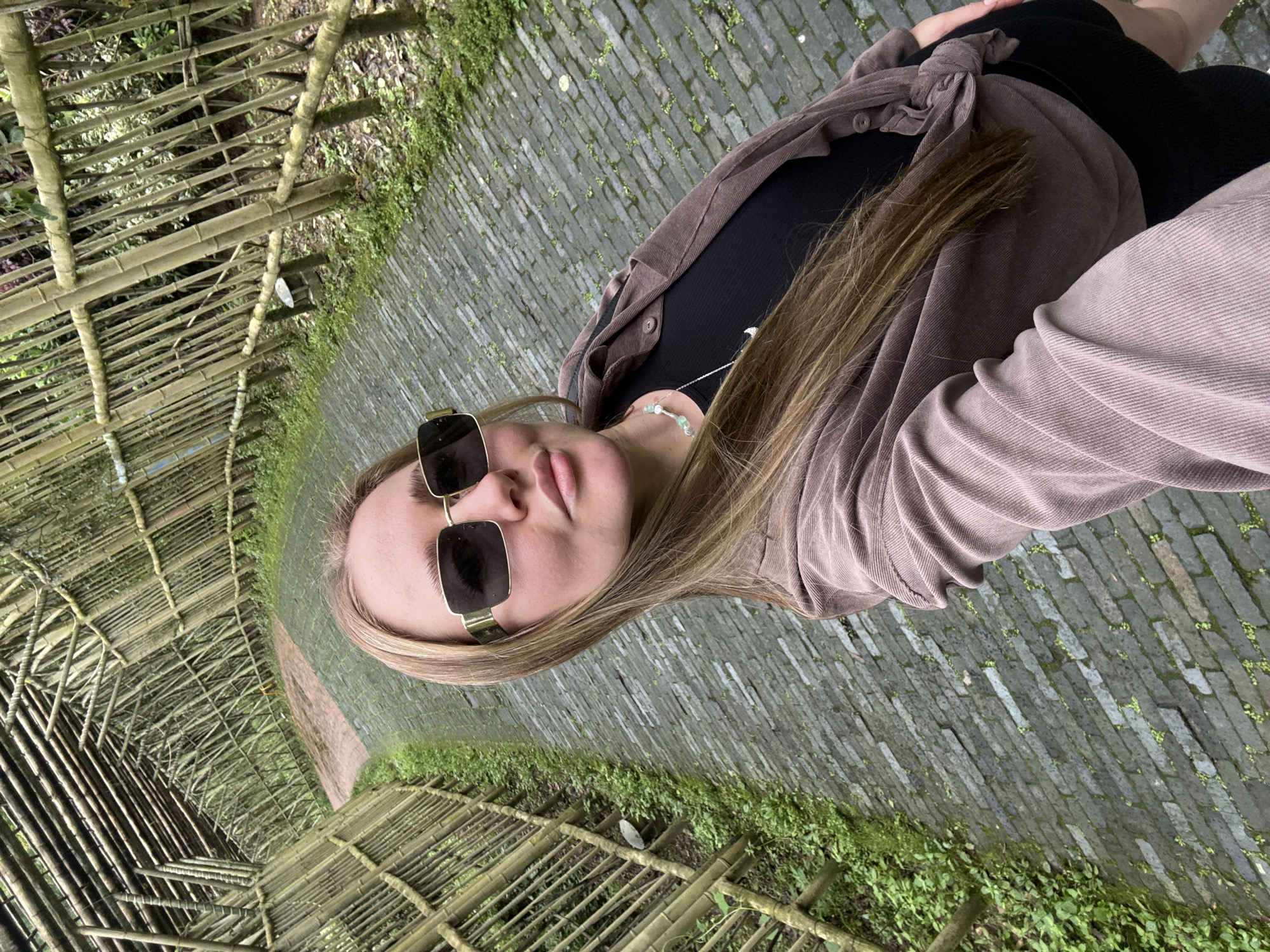 A fair-skinned woman, likely in her early to mid-twenties, is photographed in what appears to be a tourist location in Jundiping, China. She is wearing a black top, dark bottoms, a lavender corduroy shirt tied at the waist, and square sunglasses. She has long blonde hair. The location includes a brick path overgrown with patches of greenery, as well as a bamboo structure creating a tunnel-like enclosure over the path.