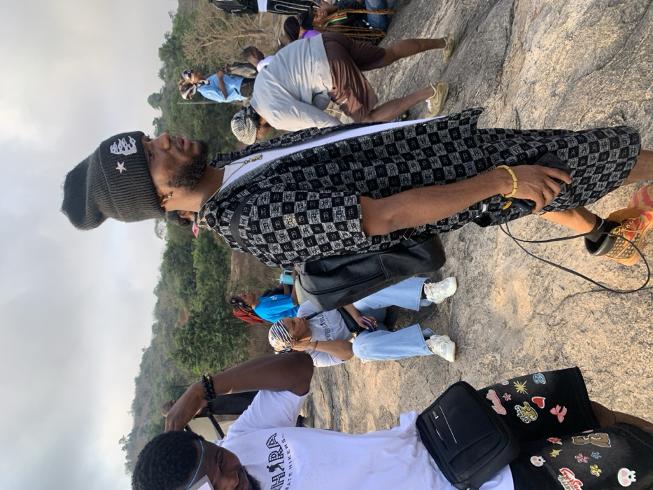 A group gathers on a rocky, elevated terrain overlooking green hills under a cloudy sky. A man in a black-and-white checkered shirt, beanie, and sneakers stands prominently. Others are seated or walking around him. There's a diversity of clothing styles, including t-shirts, jeans, and head coverings. Bags and backpacks rest on the ground, suggesting an outdoor activity like hiking or sightseeing.
