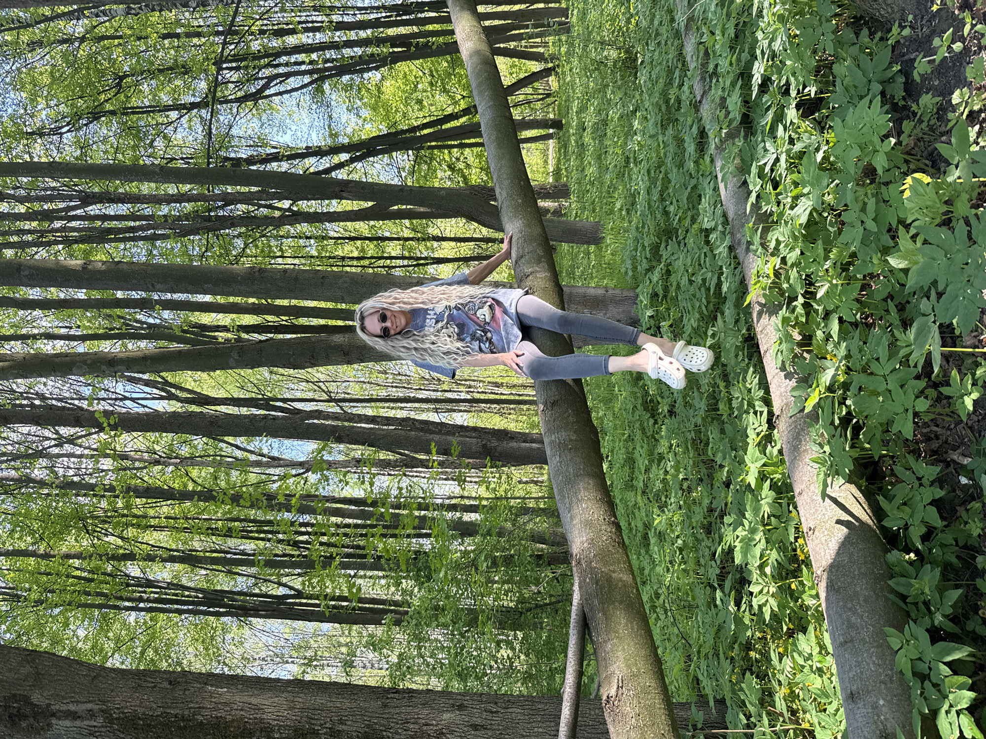 A photograph shows a woman with long blonde hair sitting on a fallen tree in a forest. She is wearing a blue t-shirt, gray pants, and white Crocs. She has sunglasses on. The surrounding forest is lush with green foliage and tall trees. The location of the photograph may be Rusanovo, Russia.