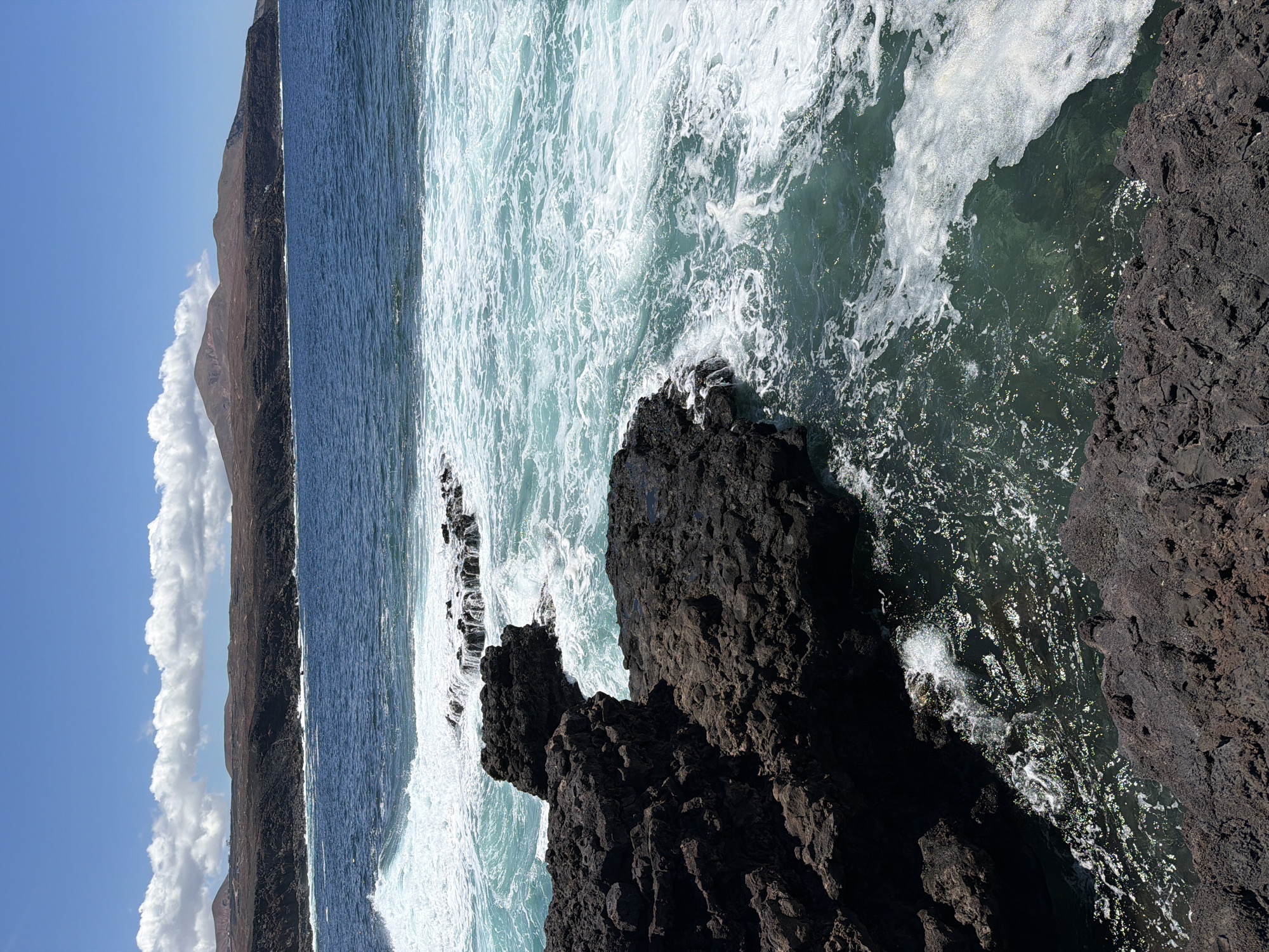 The image shows a rocky coastline with the ocean in the background, likely located in Tinajo, Spain. The foreground features dark, volcanic rock formations, while the ocean displays a mix of blue and turquoise colors with white foamy waves crashing against the rocks. A strip of land and a mountain are visible in the distance, under a blue sky with some clouds.